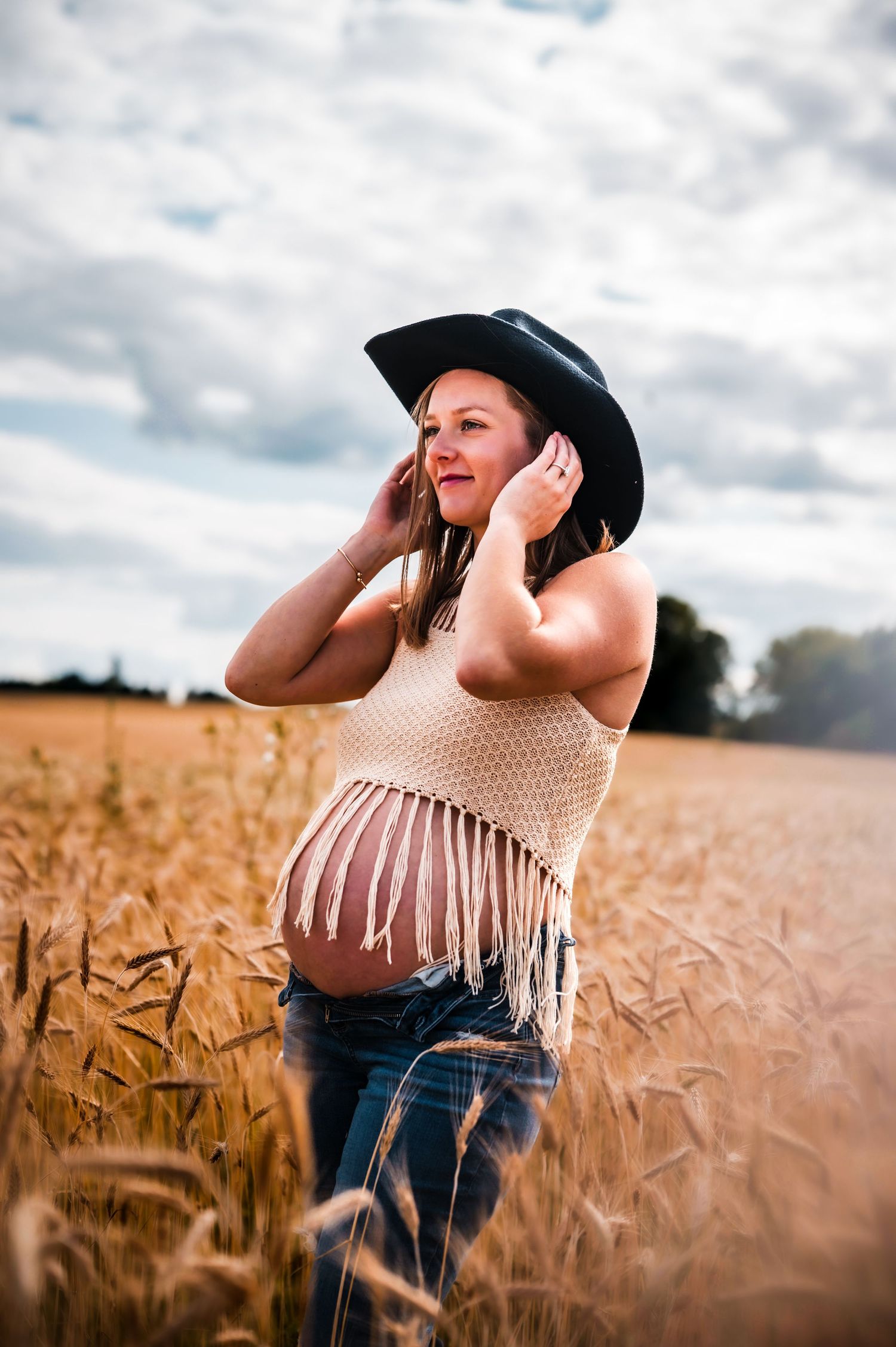 Pregnant woman in black cowboy hat and fringed crop top standing in golden wheat field under cloudy sky.
