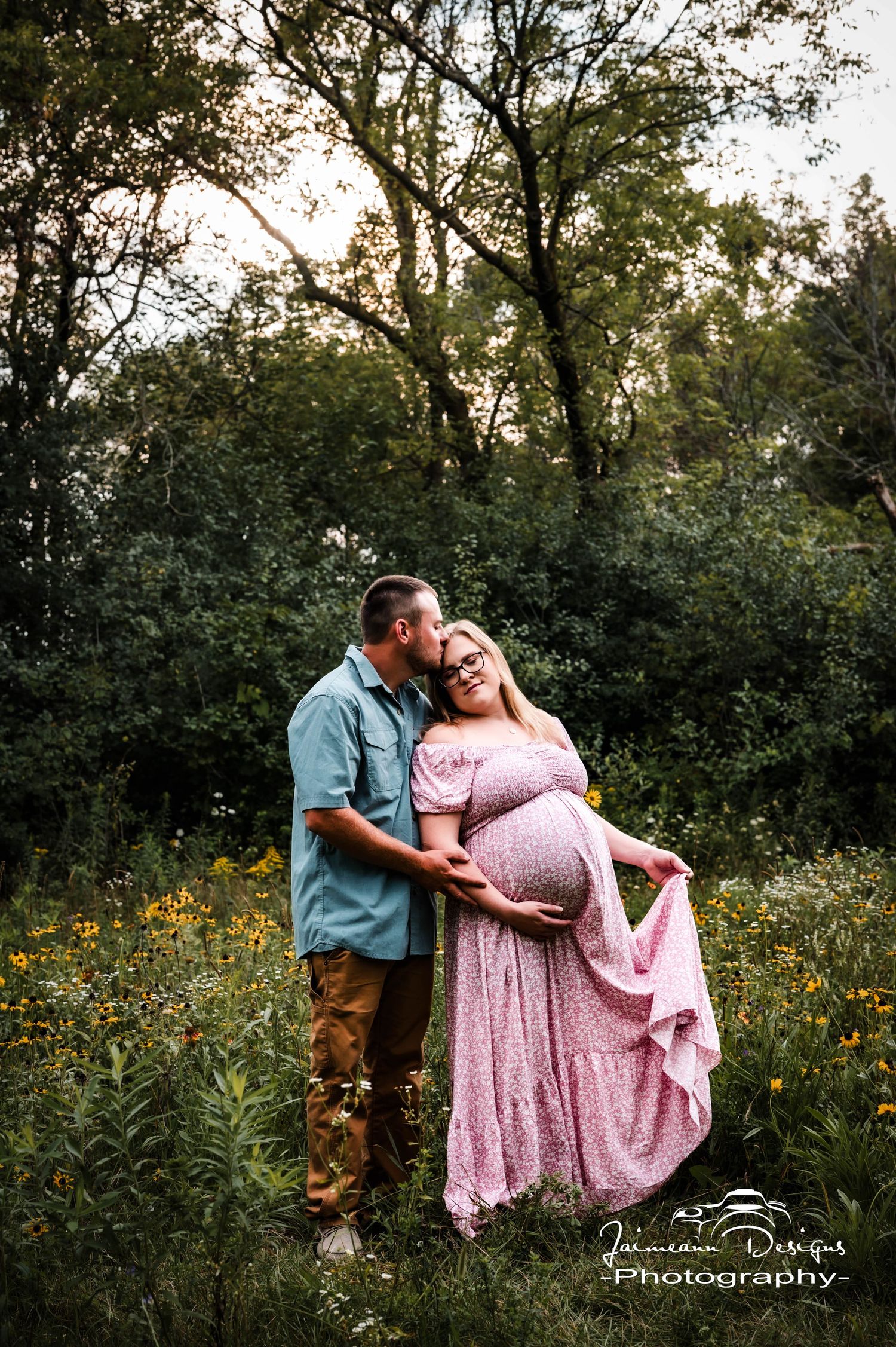 Couple poses in wildflower field, pregnant woman in pink maternity gown, partner kissing her cheek at golden hour.