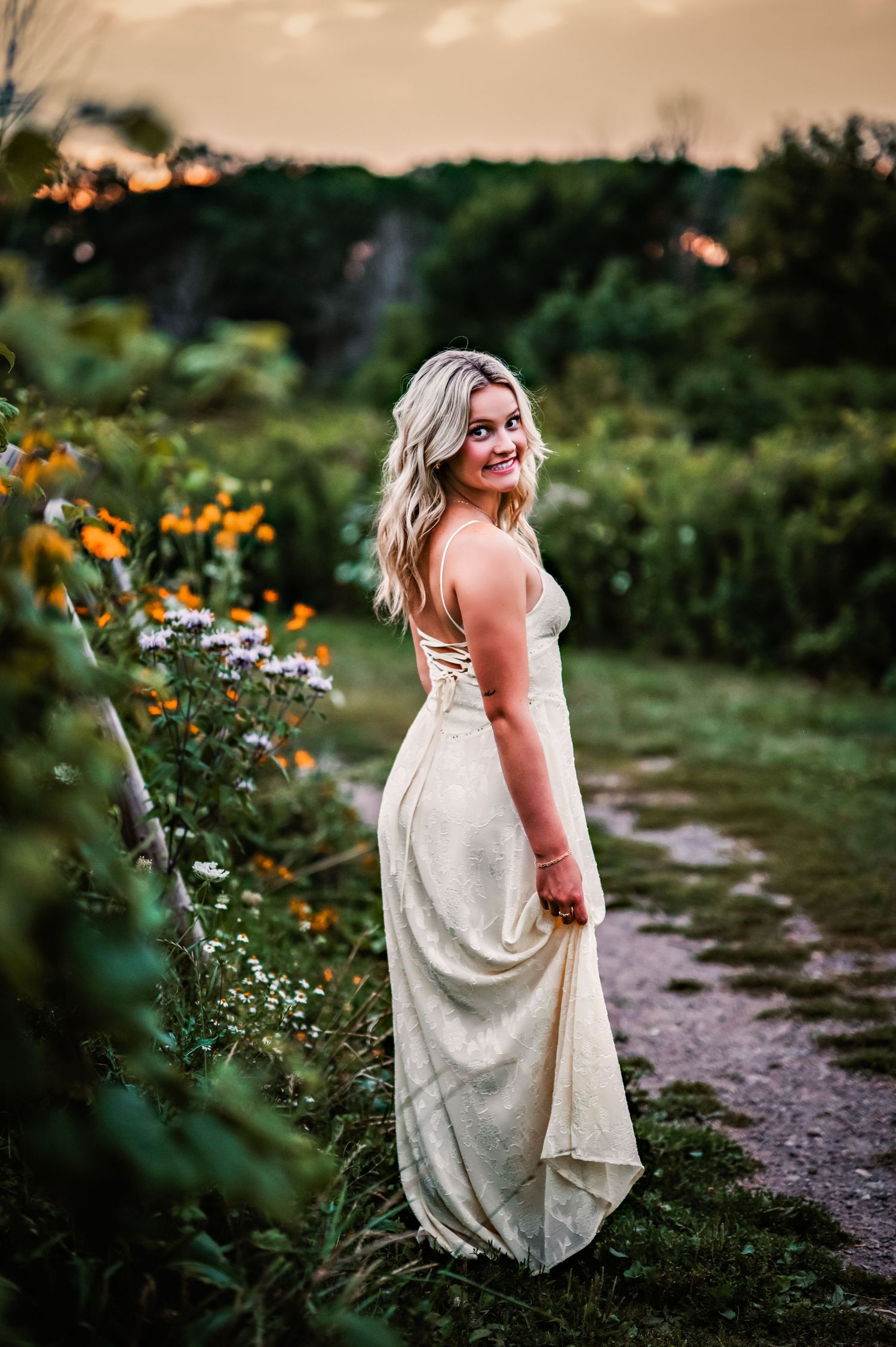 A person in a flowing white dress stands in a garden path surrounded by blooming wildflowers at sunset in Menomonee Falls Country Park, WI