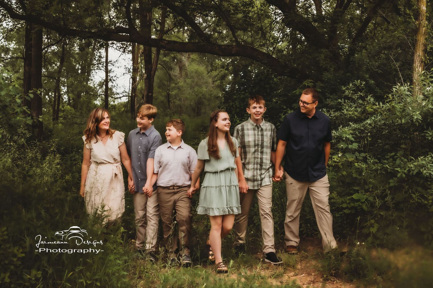 A family group poses together in coordinated casual outfits in a wooded setting.