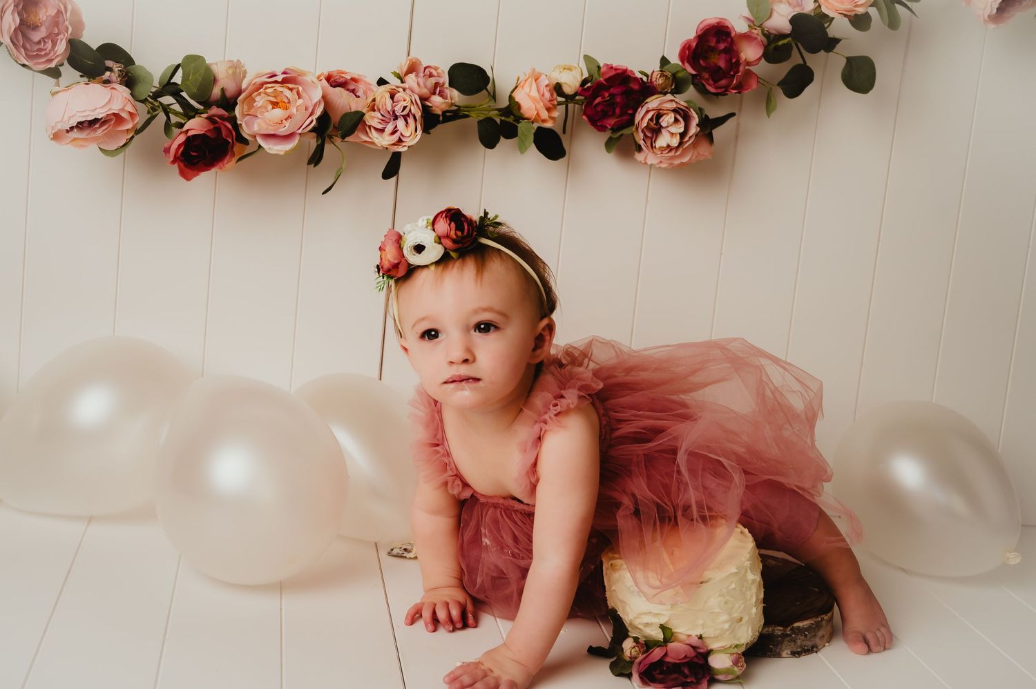 A baby wearing a dusty rose tulle dress poses beneath a romantic floral arch with white balloons.