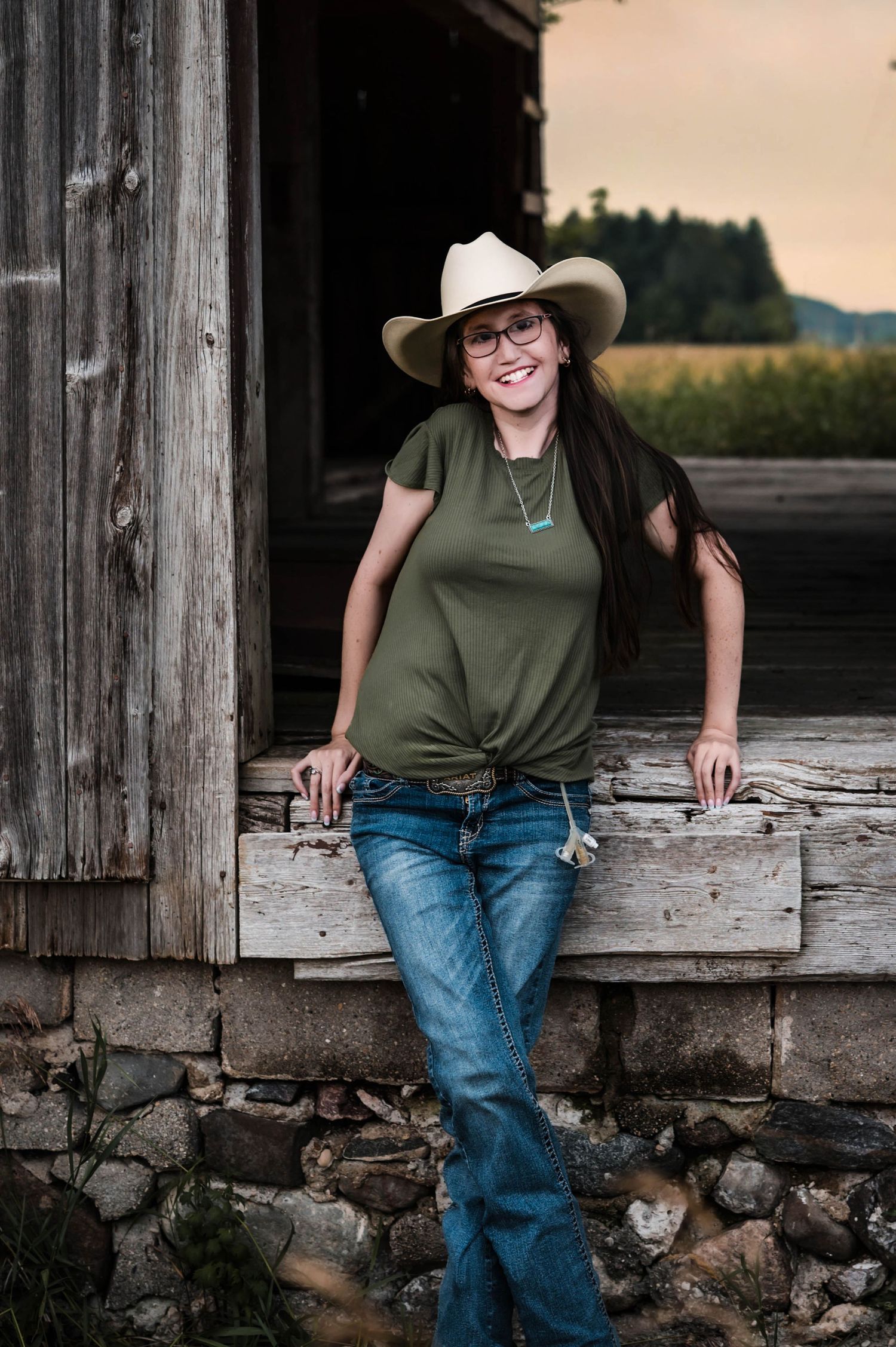 Smiling woman in cowboy hat, glasses, green shirt, and jeans leaning against rustic barn with field background.