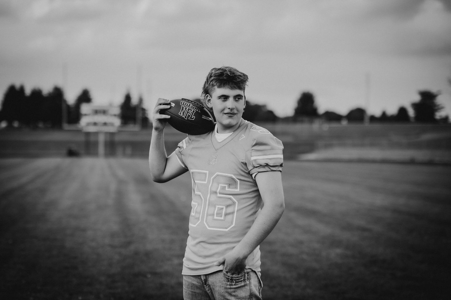 Black and white photo of a teen football player in jersey number 56 holding a helmet on a field.