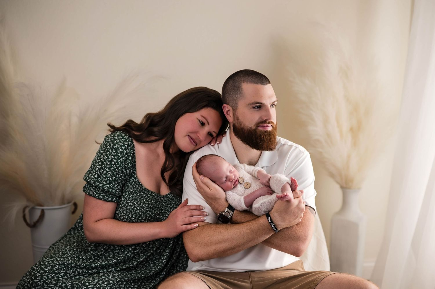 Smiling couple sitting together in a bright studio cradling their sleeping newborn baby, with soft pampas grass decor in the background.