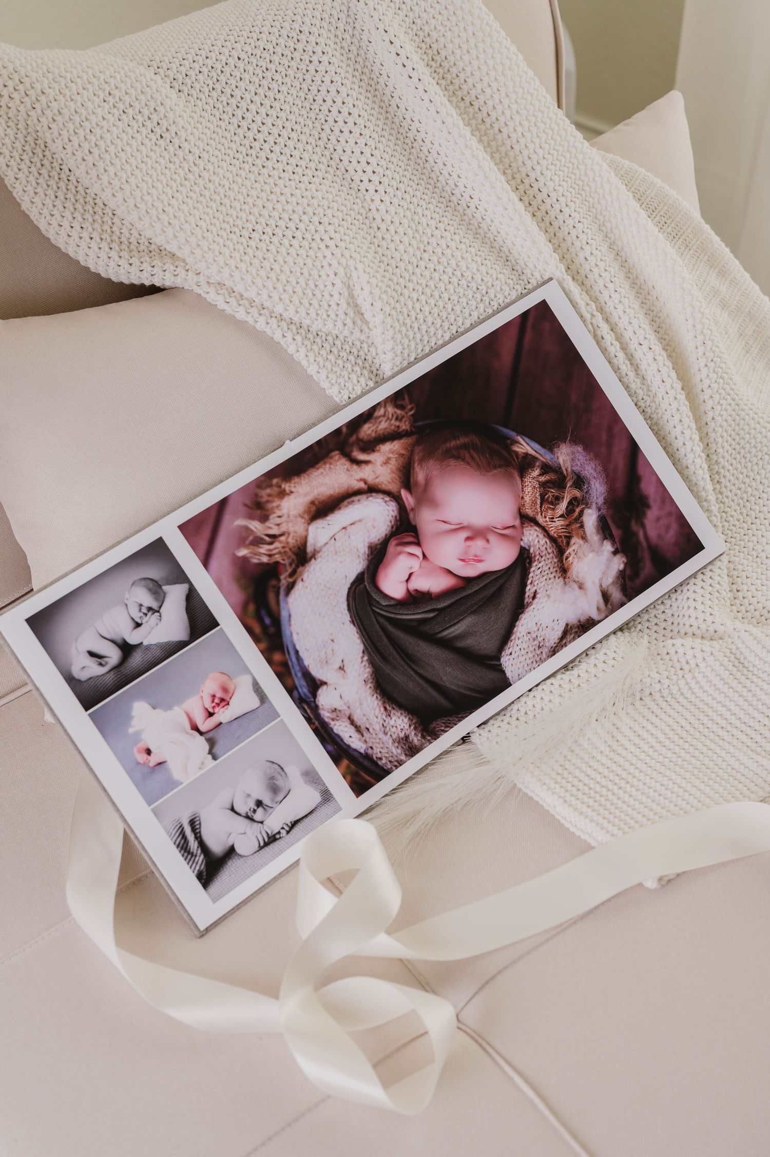 Newborn photography prints and photo book displayed on cream leather sofa with white knit blanket, featuring sleeping baby portraits.