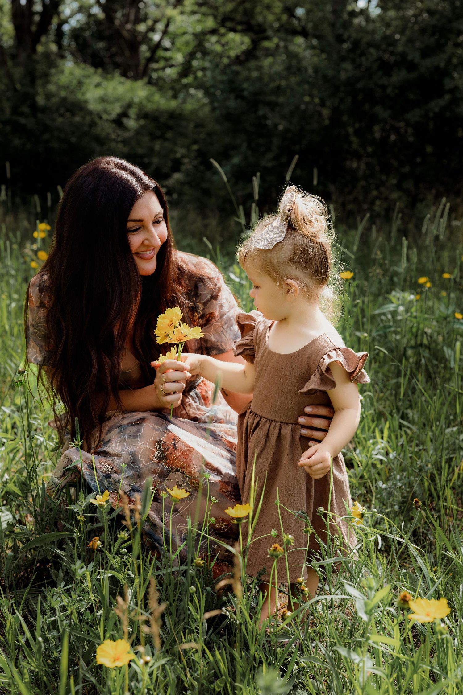 A mother and toddler girl share a tender moment with yellow wildflowers in a lush green meadow.