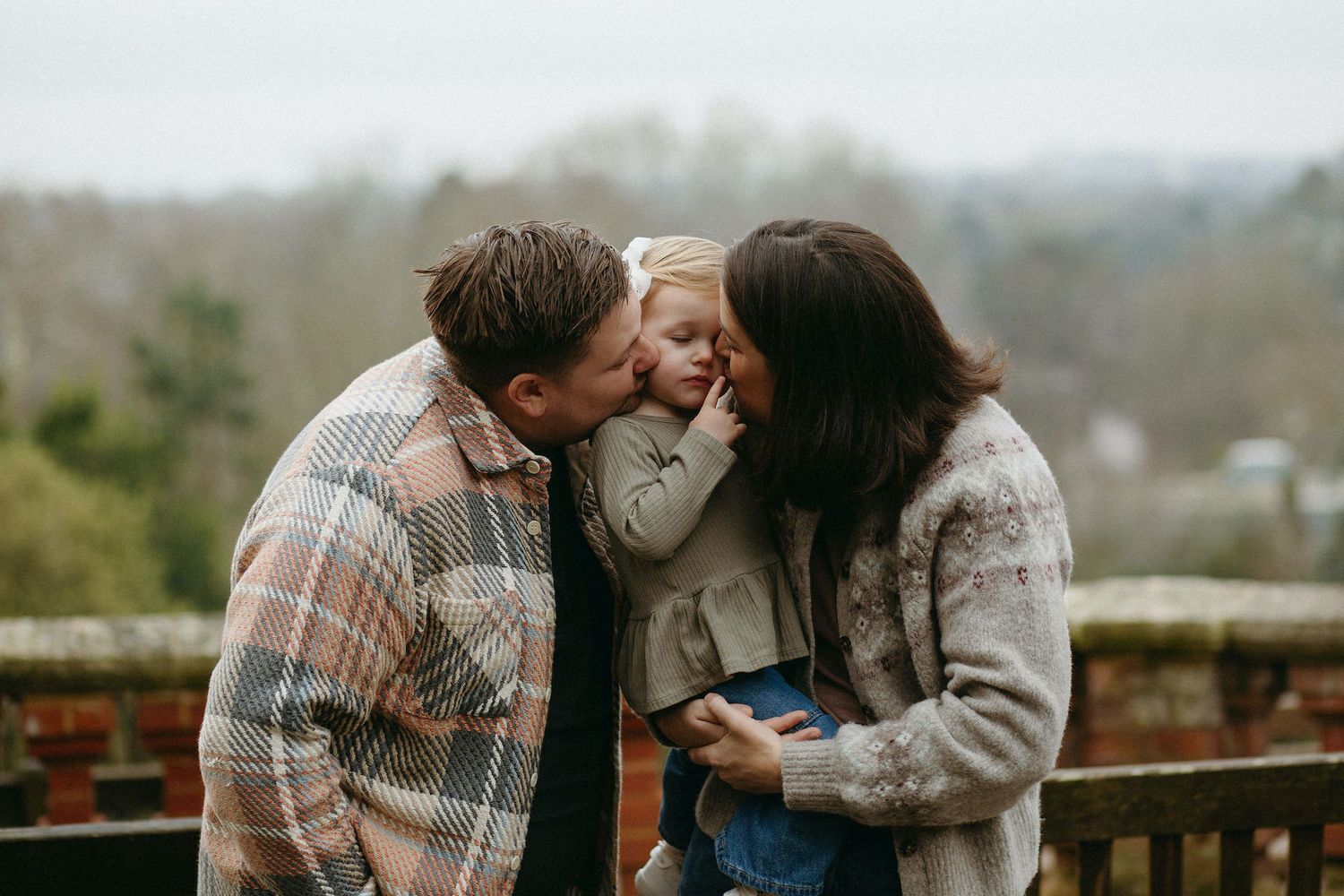 Parents kissing their toddler on the cheeks outdoors, sharing a tender family moment with lush greenery in the background.