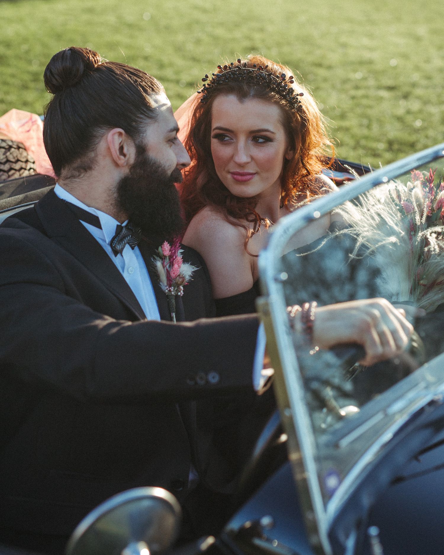 A bearded groom in a black tuxedo and a redheaded bride in a vintage convertible car share a romantic glance.