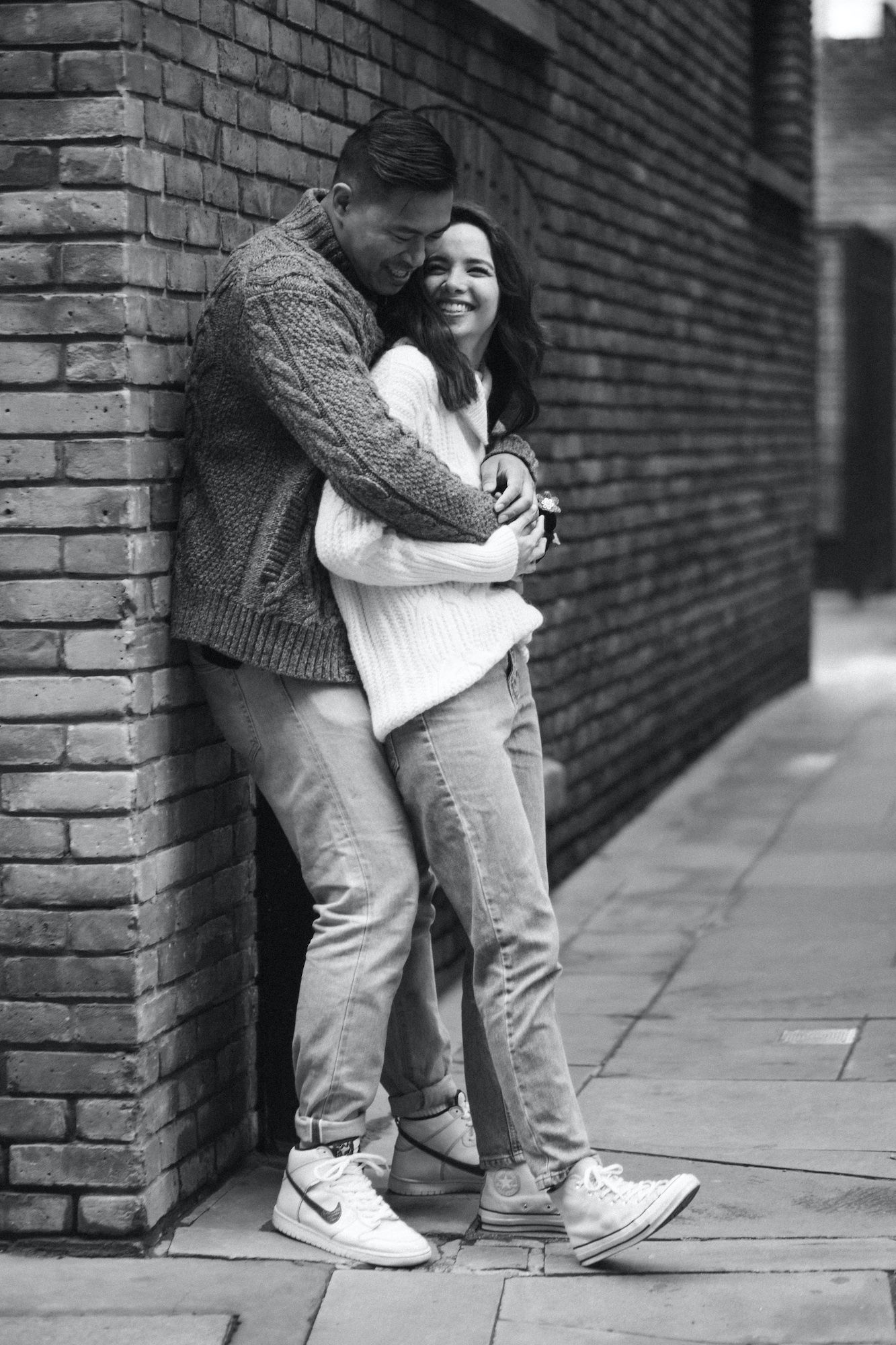 Black and white photo of a couple embracing against a brick wall, woman smiling joyfully in casual jeans and sweaters.