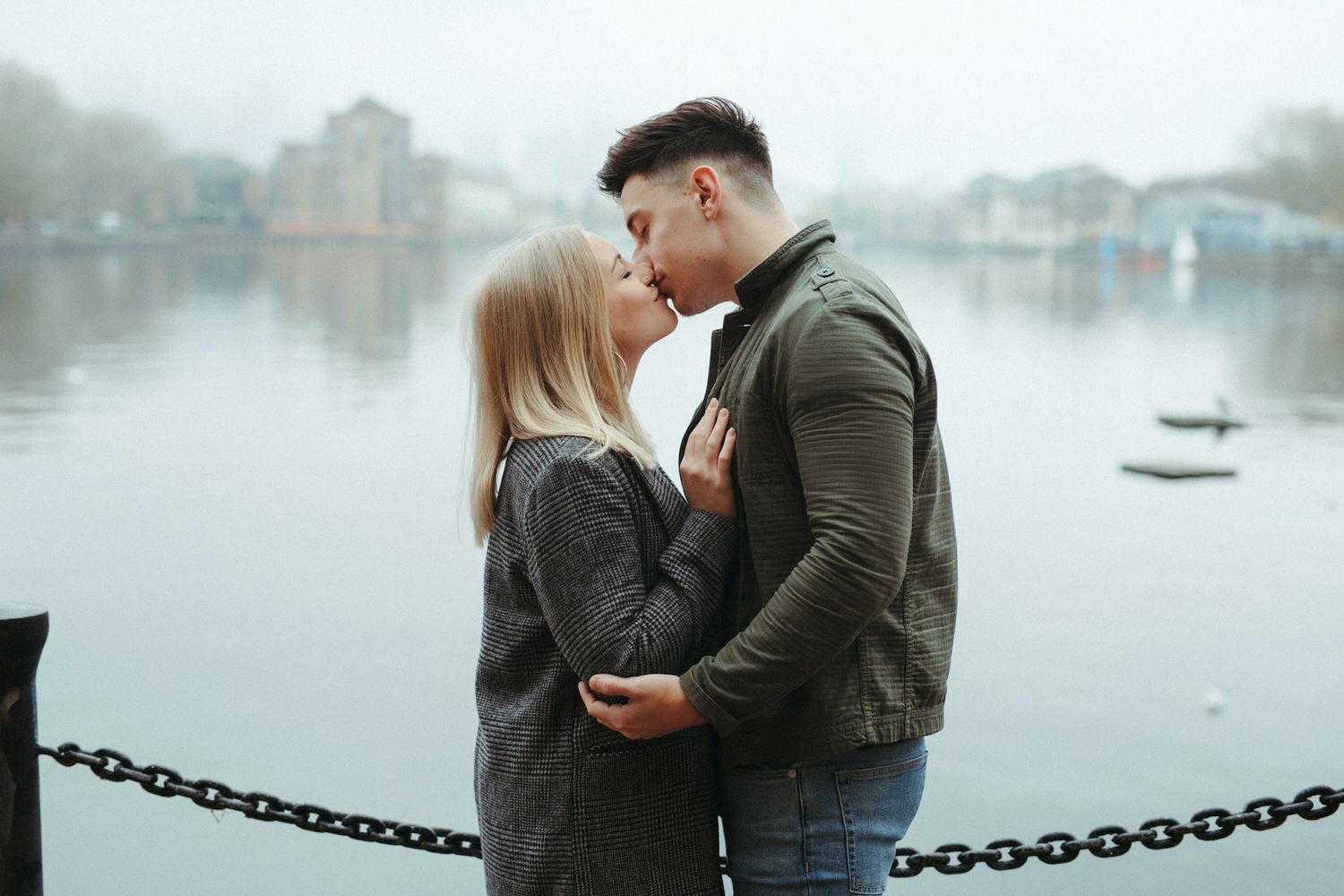 A young couple shares a romantic kiss by a misty lakeside, dressed in casual autumn outfits.