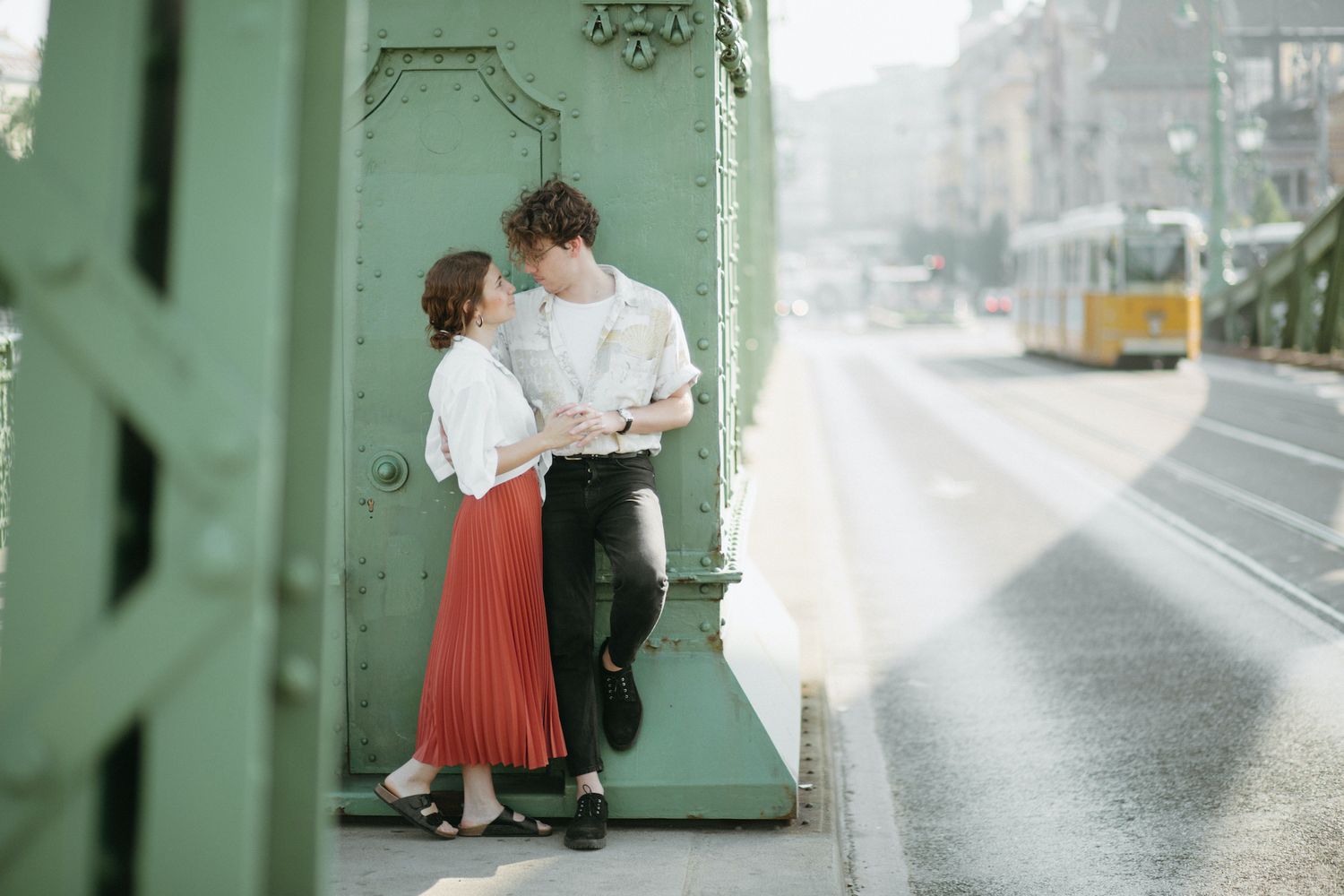 A romantic couple stands close together on a green bridge, with a yellow tram passing in the background.