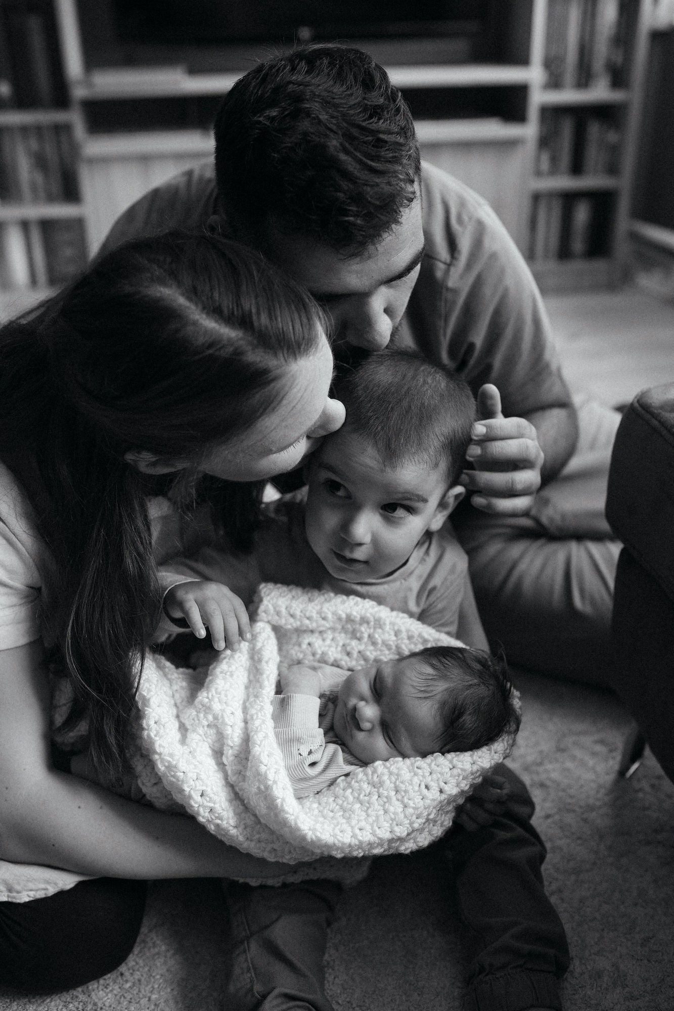 Black and white family photo of parents kissing toddler while holding swaddled newborn baby at home.