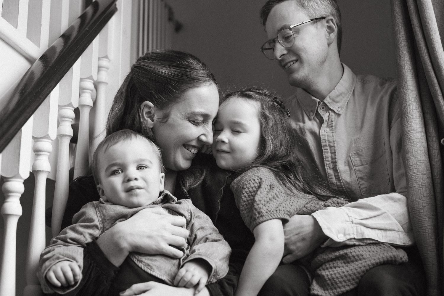Black and white family portrait of parents and two young children sitting together on staircase, smiling warmly.