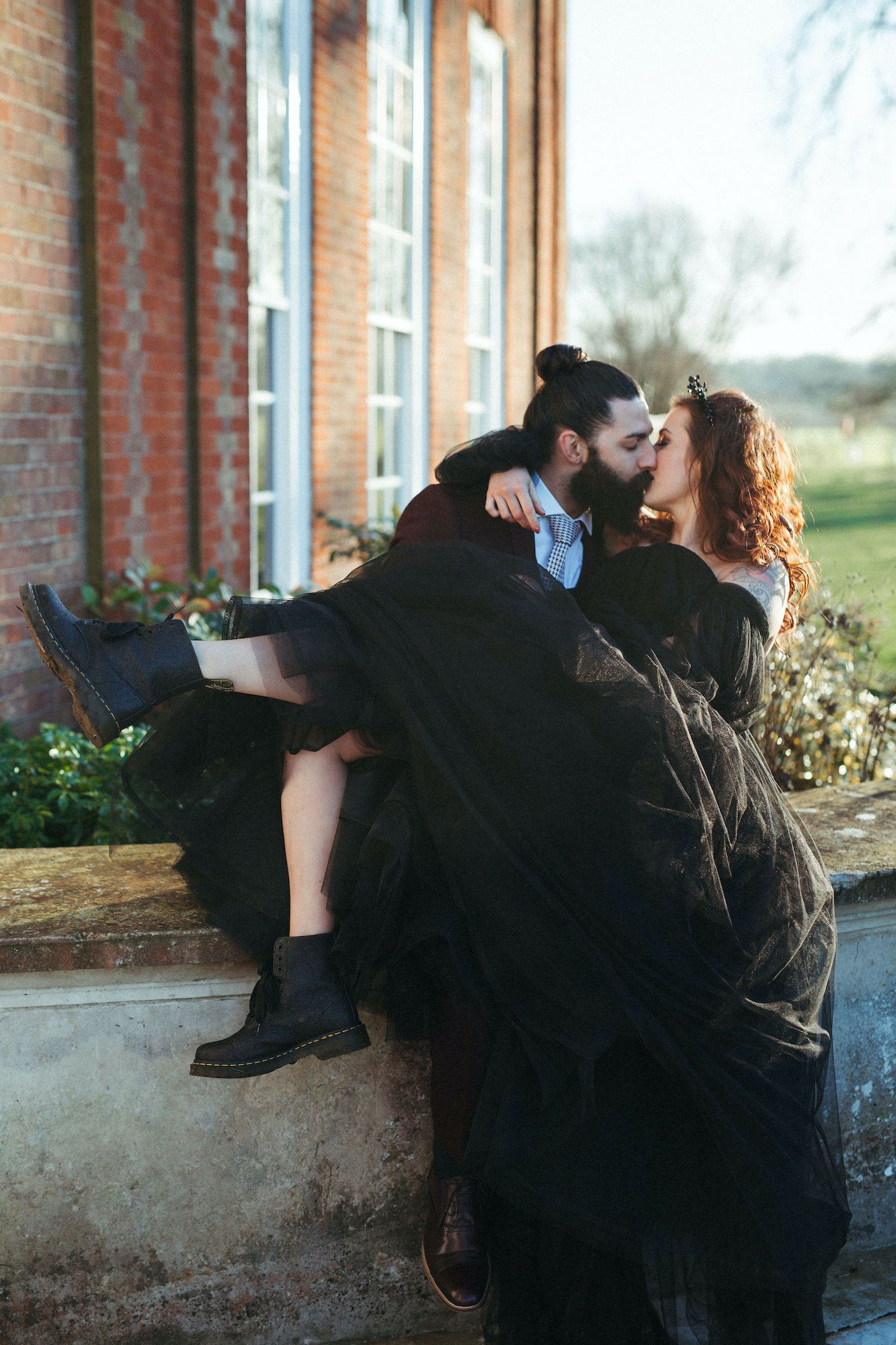 A couple in black wedding attire share a romantic kiss outside a brick building, with the groom holding the bride.