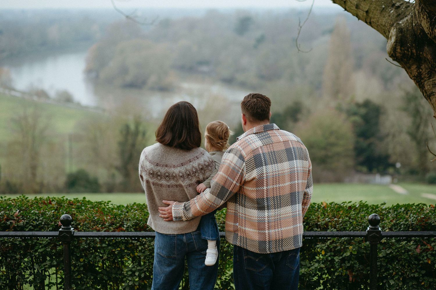 Family of three standing together overlooking a scenic river and green landscape on a cloudy day.