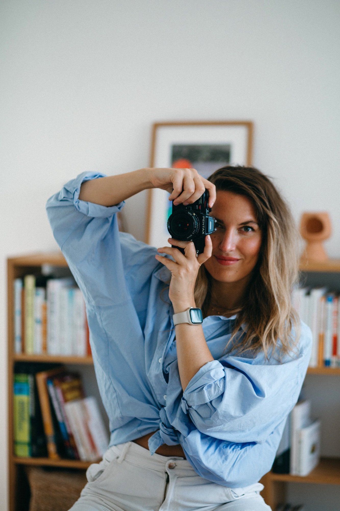 Woman in blue shirt holds analogue camera up to eye, posing in front of bookshelf in bright modern interior.