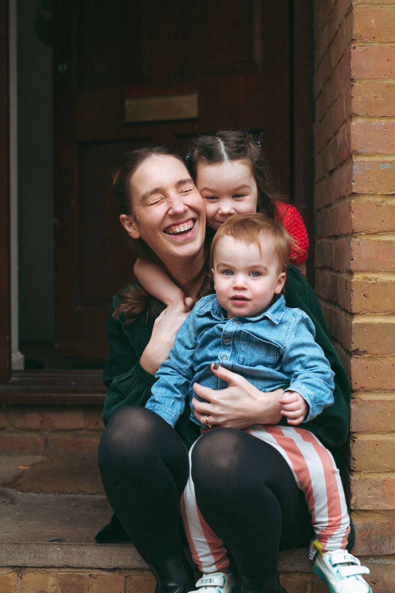 A laughing mother sits on steps holding a baby in denim, while an older child hugs her from behind.