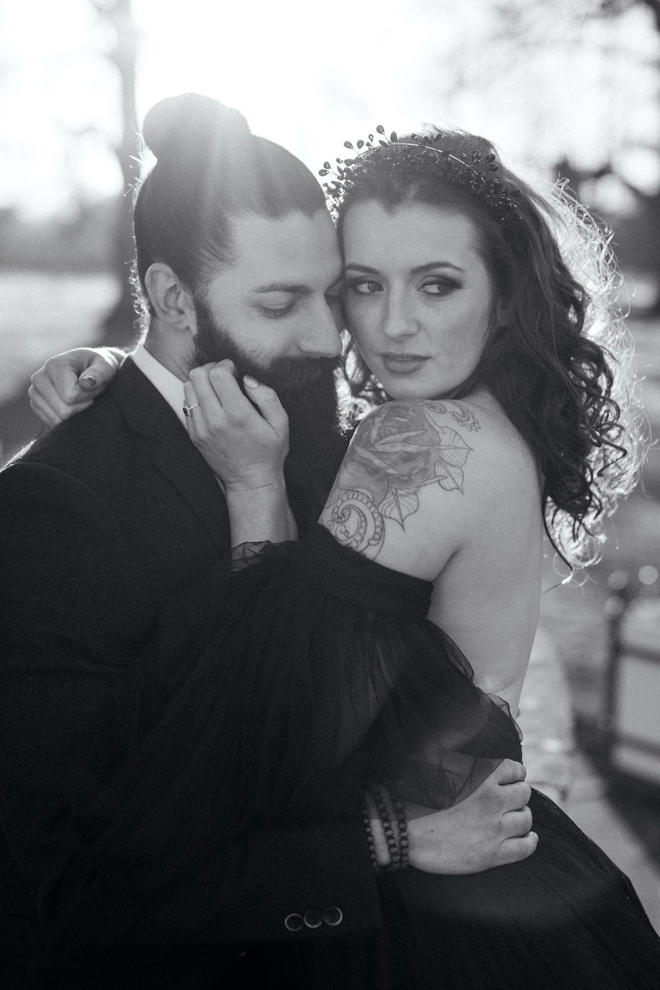 Black and white wedding photo of bearded groom embracing tattooed bride in dark gown with floral shoulder tattoo.