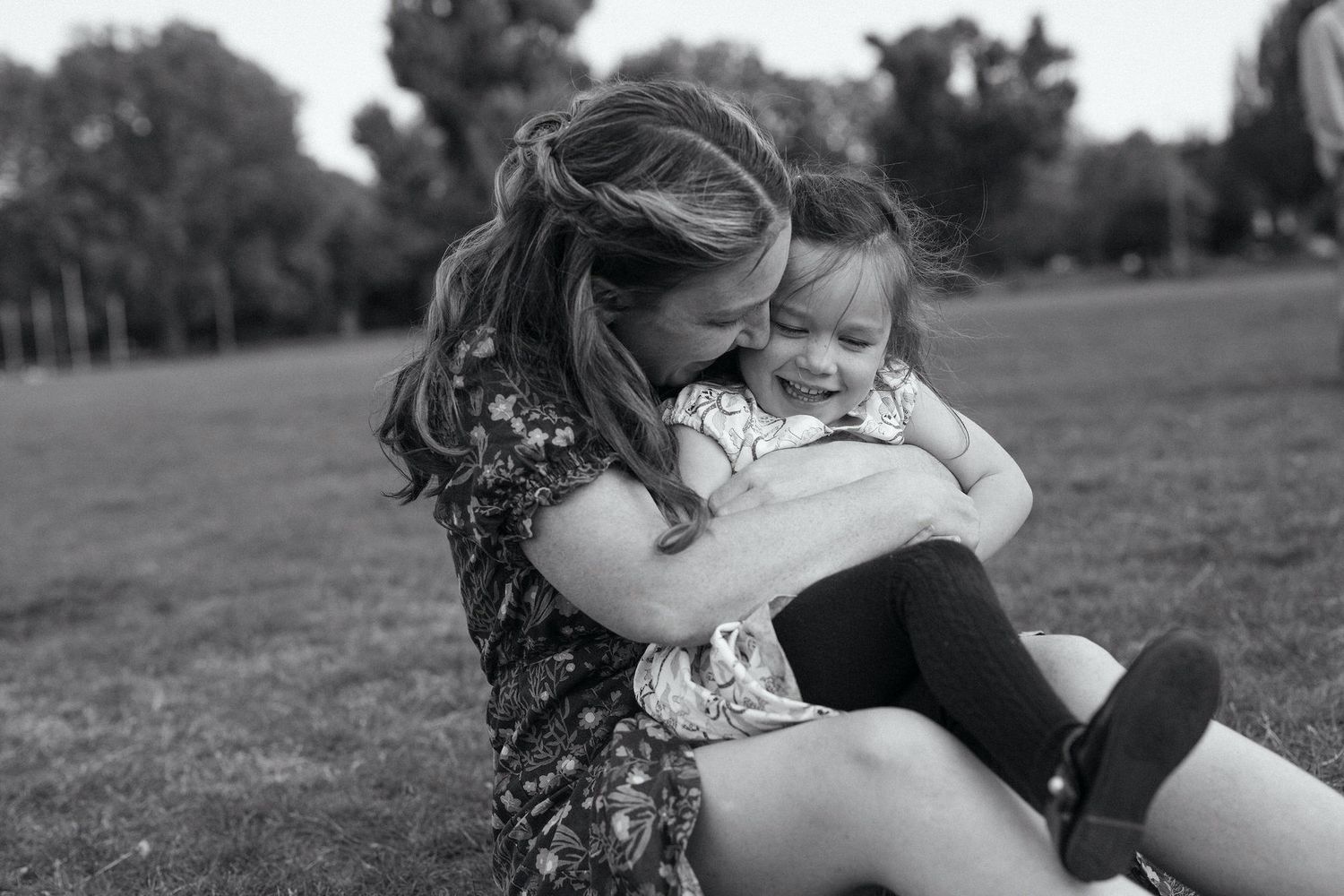Black and white photo of a mother hugging and kissing her young daughter outdoors in a park.