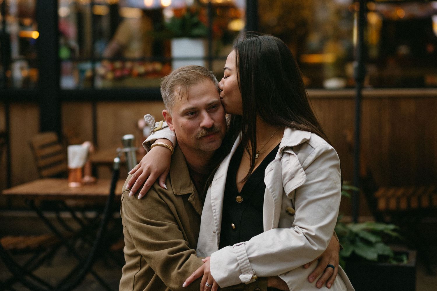 A woman kisses a smiling man on the cheek while embracing outdoors at a cozy restaurant patio.