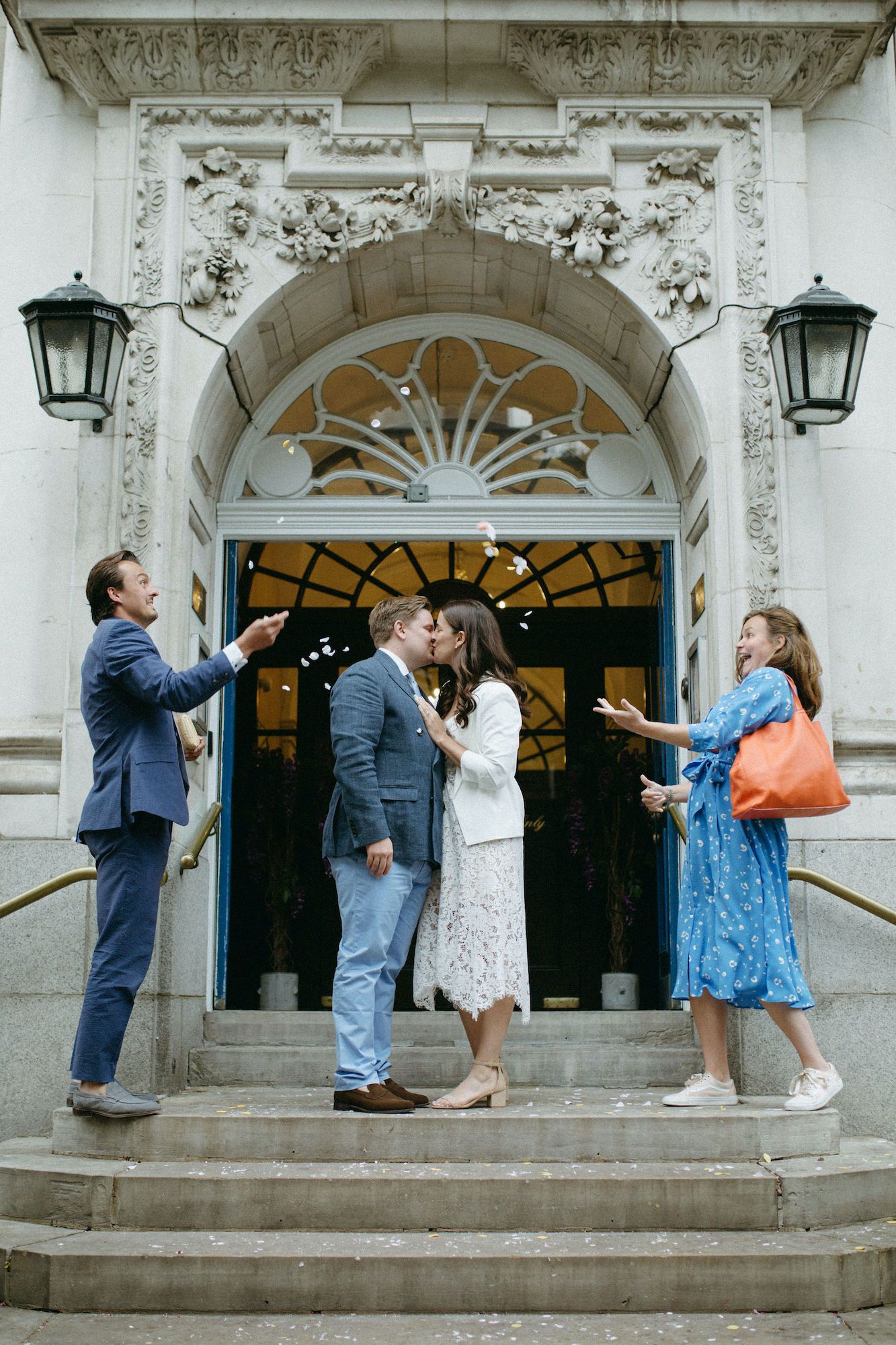 Couple sharing a kiss on ornate building steps while two guests toss confetti during an intimate city hall wedding ceremony.