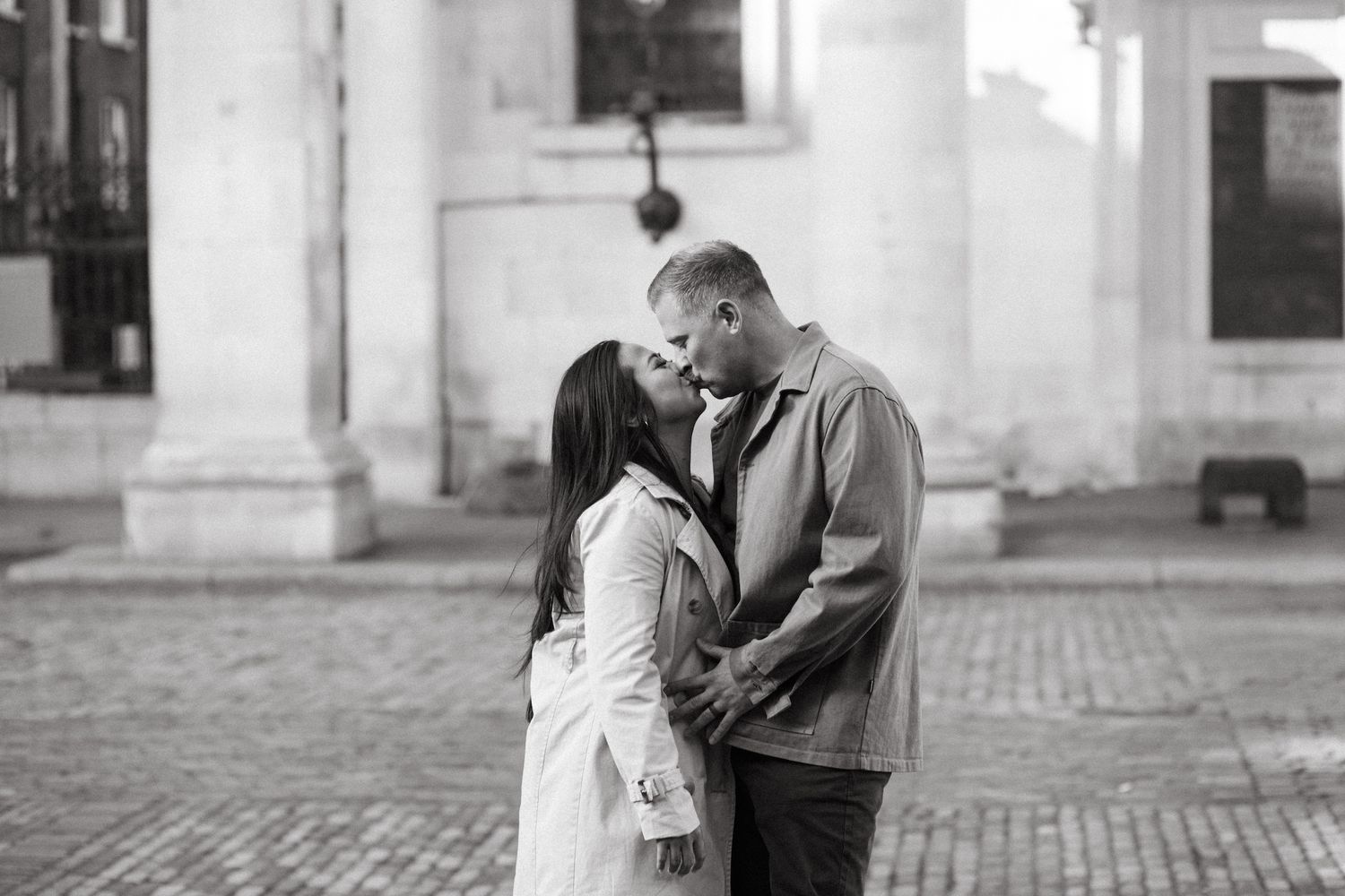 Black and white photo of a couple kissing tenderly while holding hands on a cobblestone plaza.