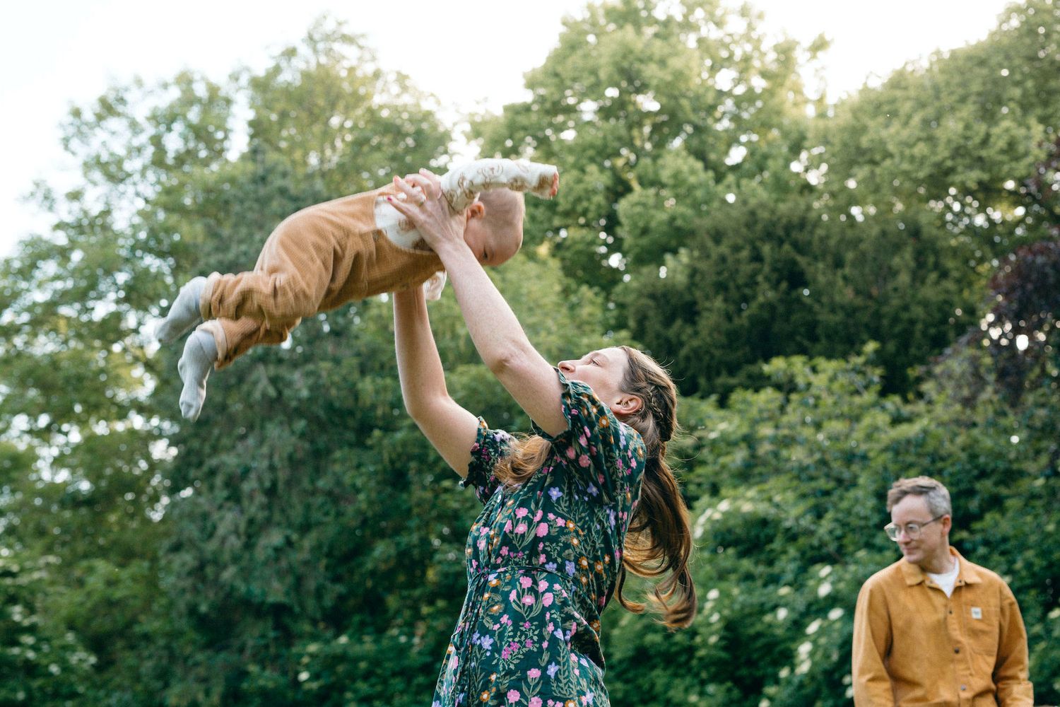 A joyful mother lifts her baby in the air outdoors, with the smiling father and lush green trees in the background.