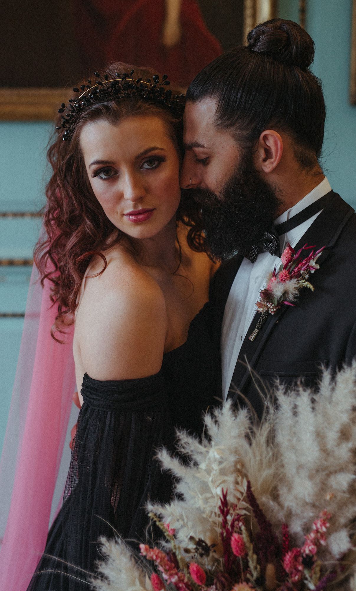 Gothic bride in black gown with pink veil and dark crown poses with bearded groom in tuxedo holding dried floral bouquet.