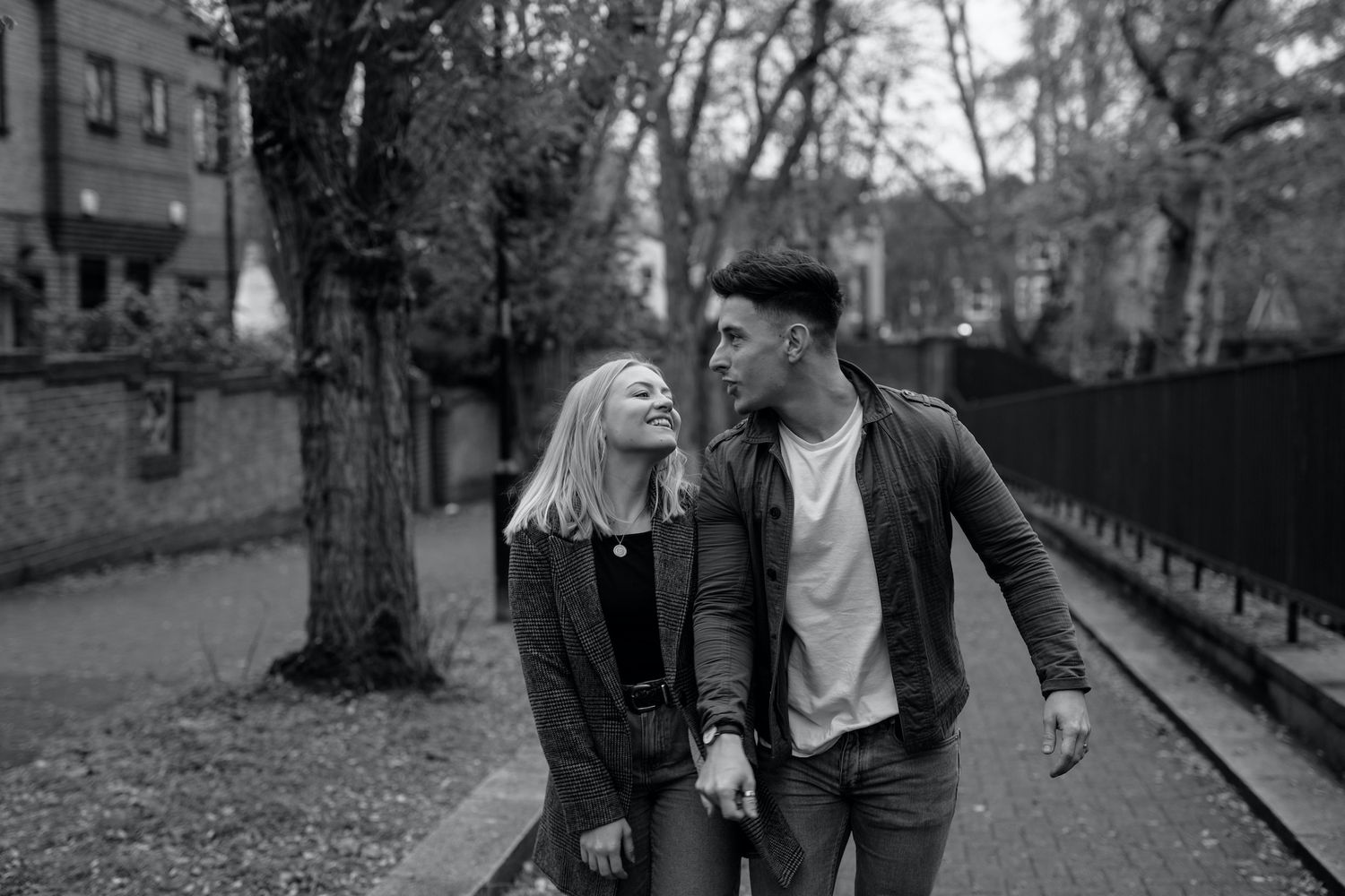 Black and white photo of a young couple walking and smiling together on a tree-lined urban pathway.