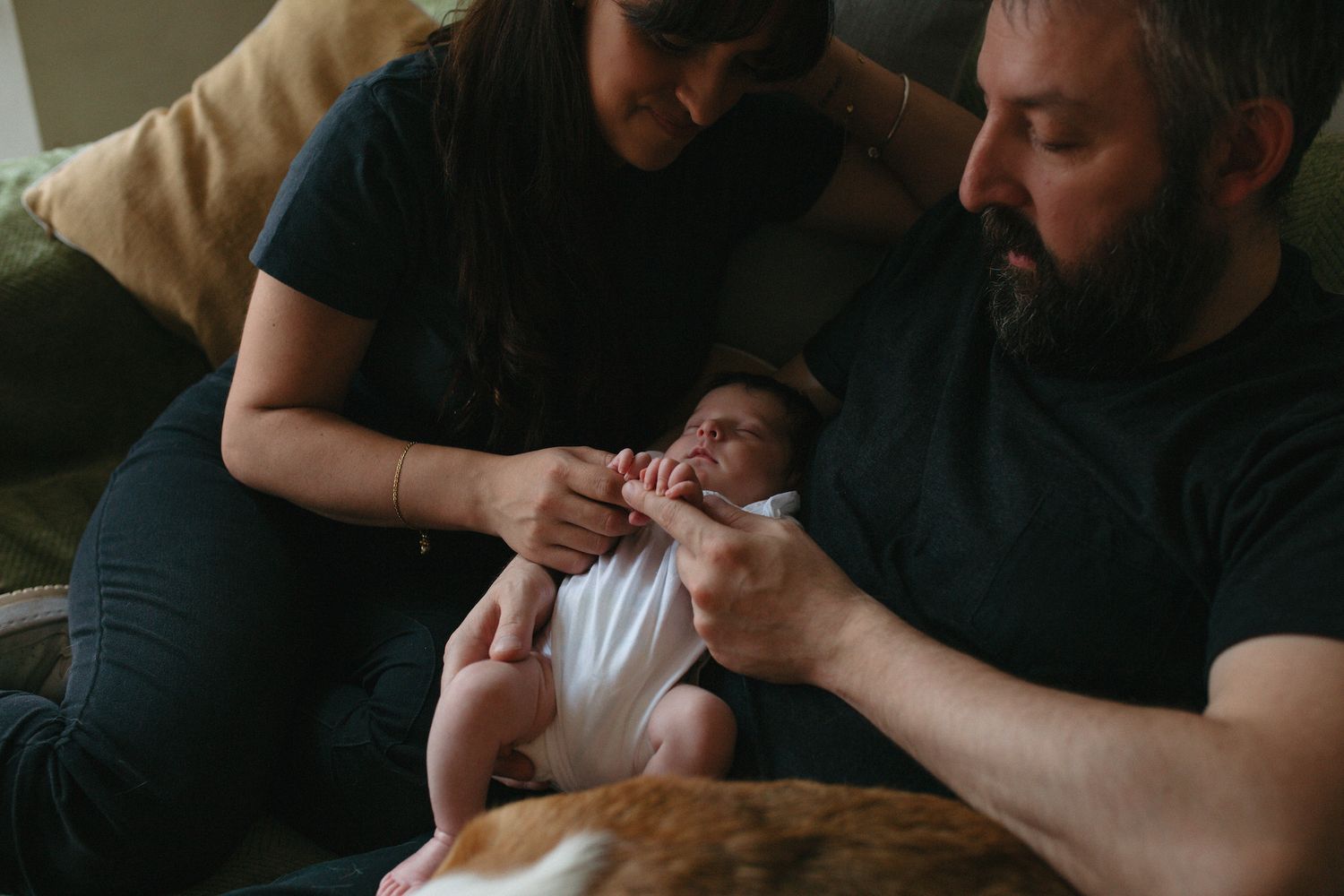 Parents lovingly holding their newborn baby on a couch with a dog nearby in a warm home setting.