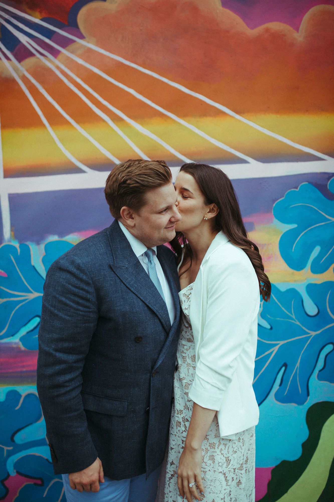 Couple sharing a romantic kiss in front of a vibrant colorful mural, woman in white dress and man in gray suit.