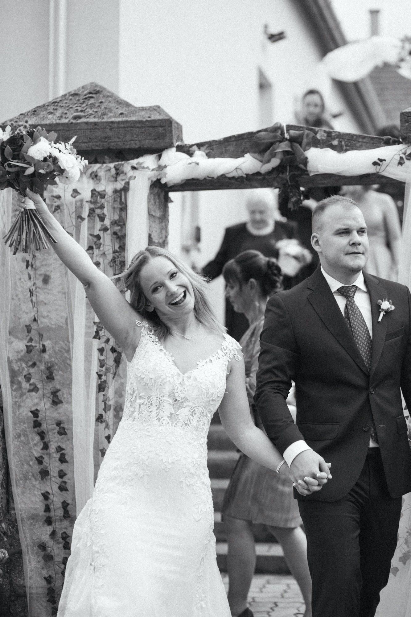 Black and white photo of a joyful bride raising bouquet while holding hands with groom after wedding ceremony.