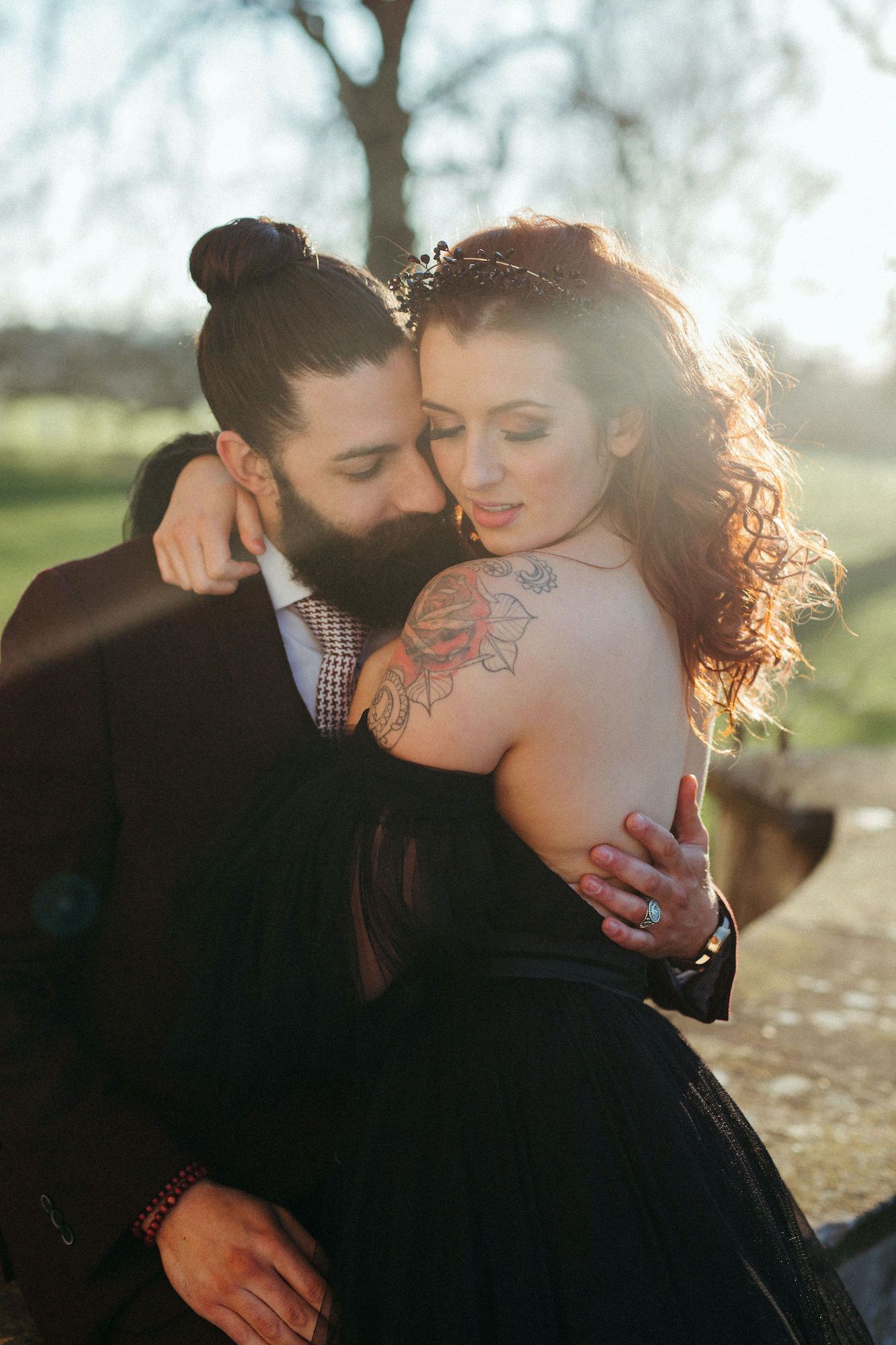 A bearded groom in a dark suit embraces a tattooed bride in a black dress, backlit by golden sunlight outdoors.