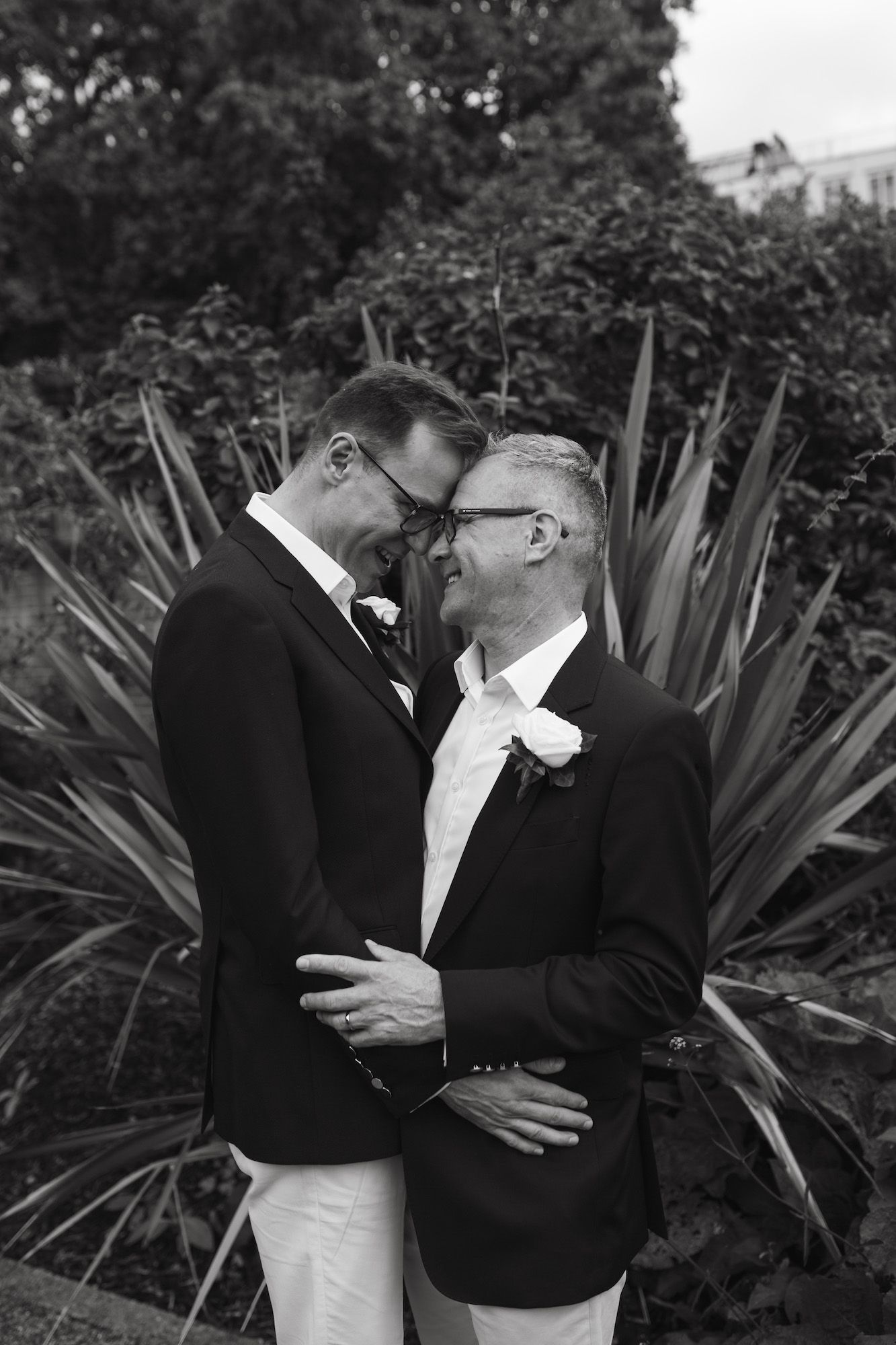 Black and white photo of two grooms in blazers embracing and smiling at each other outdoors on their wedding day.