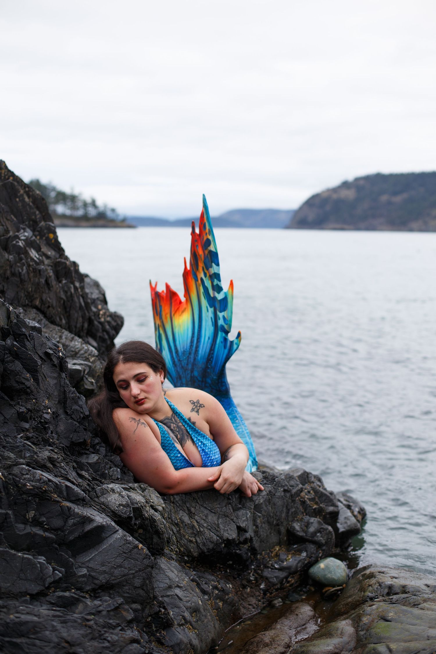 A photo of a mermaid with a vibrant tail laying on obsidian black rocks.
