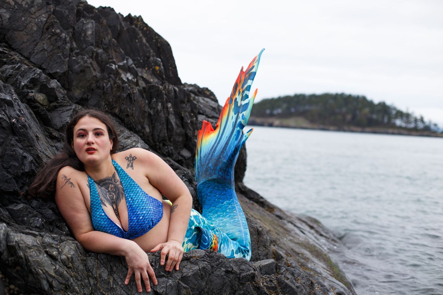 A photo of a mermaid with a vibrant tail laying on obsidian black rocks.