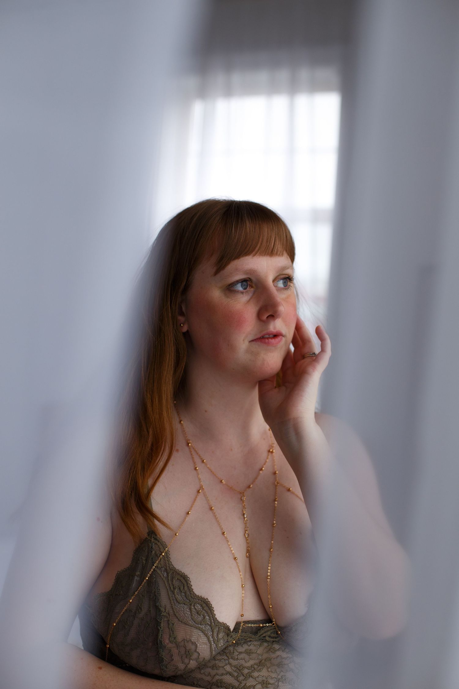A boudoir photo of woman in green lingerie, framed between sheer curtains, looking out a window