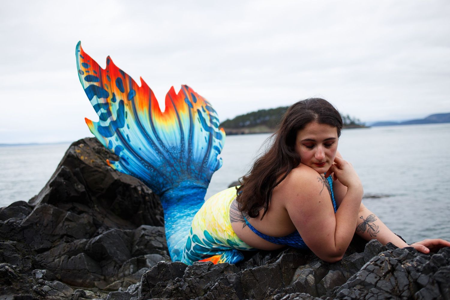 A photo of a mermaid with a vibrant tail laying on obsidian black rocks with the ocean behind her.