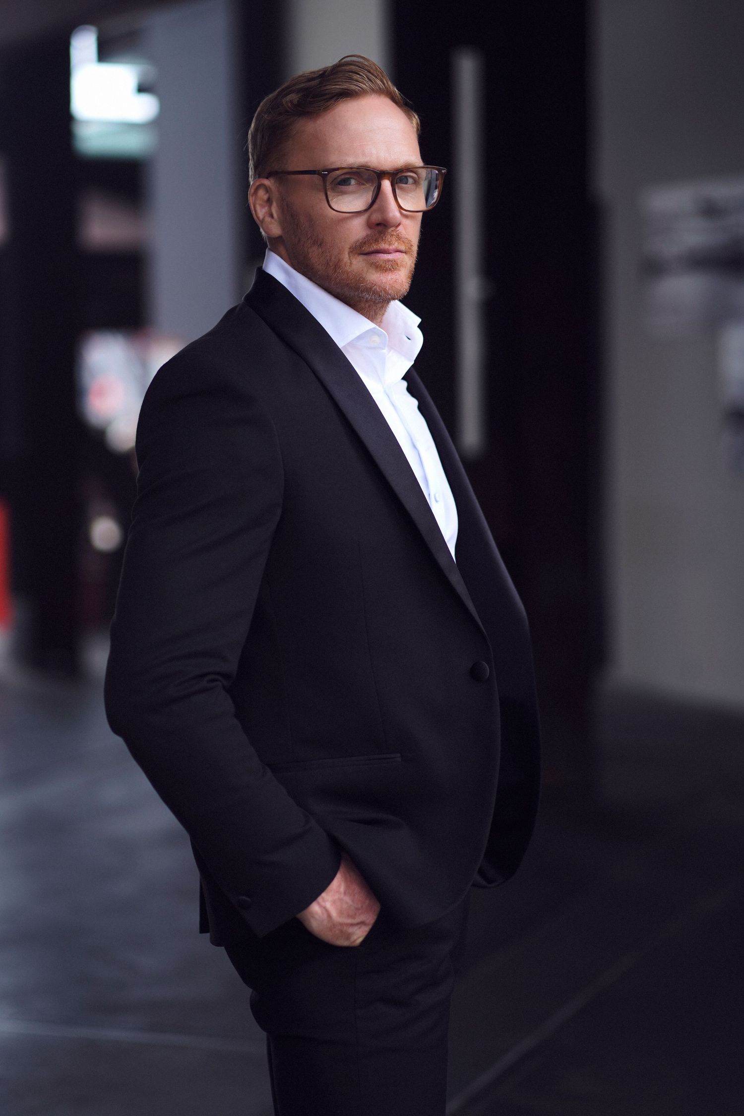 A man in glasses wearing a formal black suit and white shirt poses confidently in an urban setting at night. Studio TingTing.
