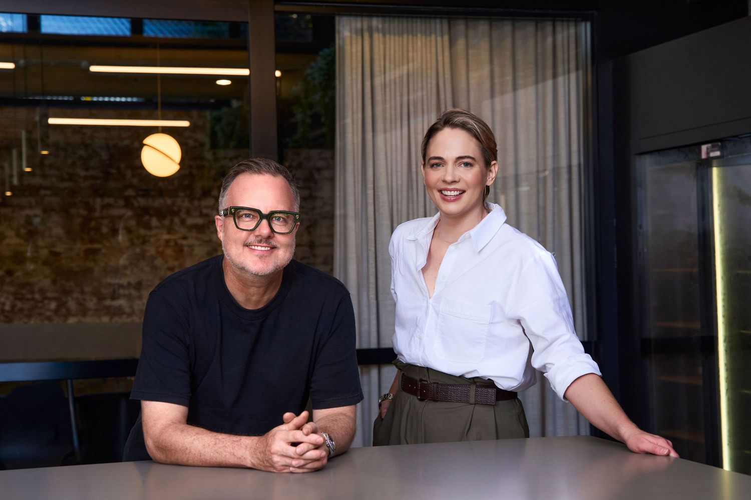 Two professionals smiling together in a modern office, man in black shirt and woman in white blouse posing at a table.