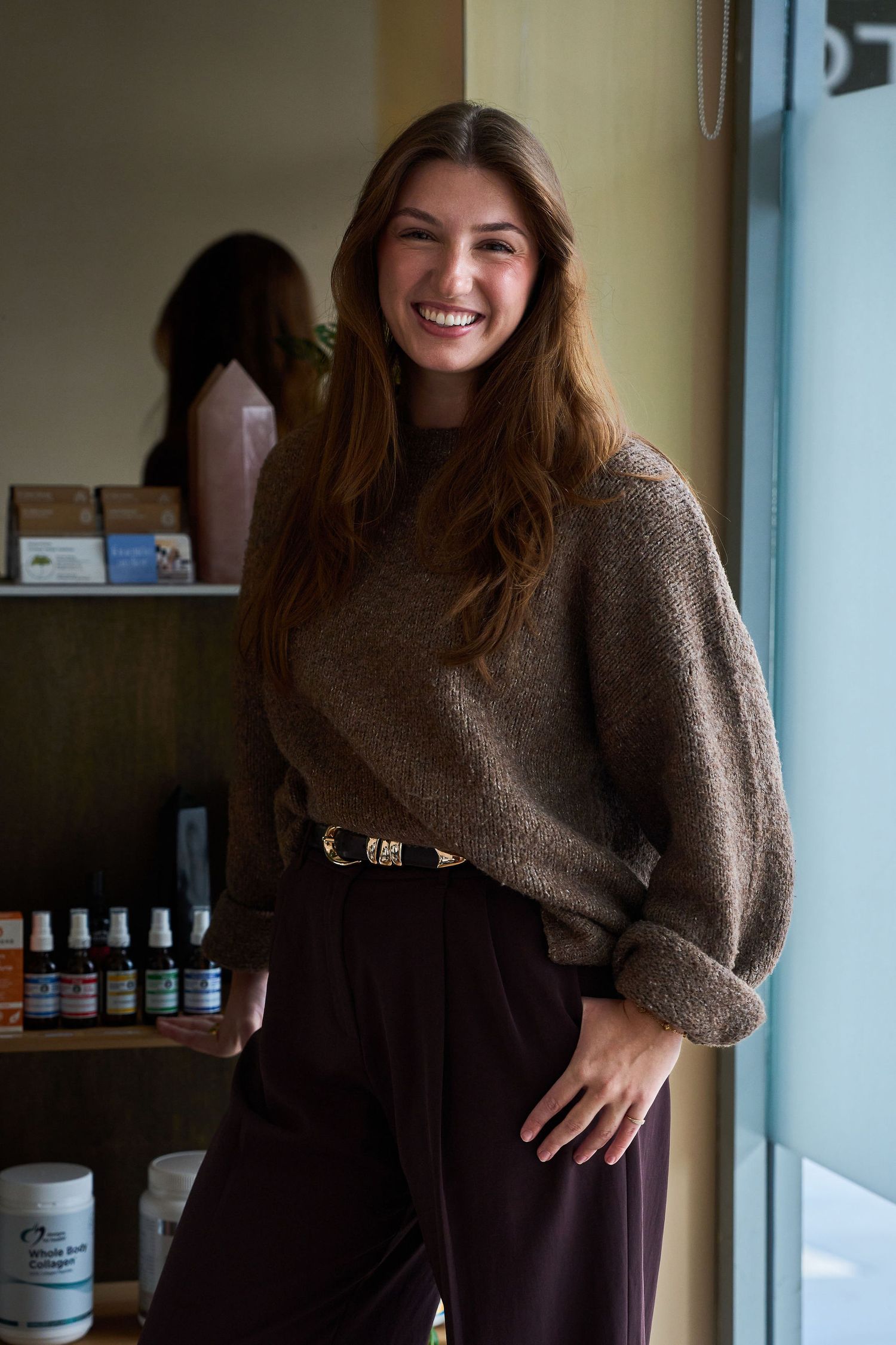 Smiling young woman in brown sweater and dark pants standing in a wellness shop with products on shelves behind her.