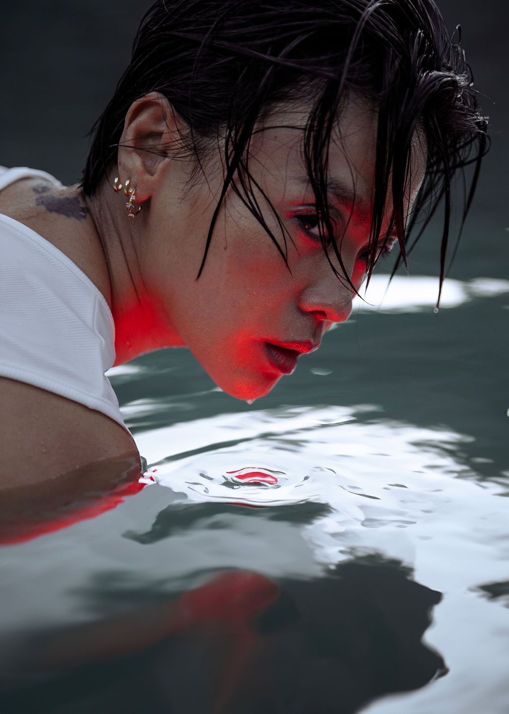 Person with wet hair and tattoos submerged in water, illuminated by dramatic red light, wearing a white top.