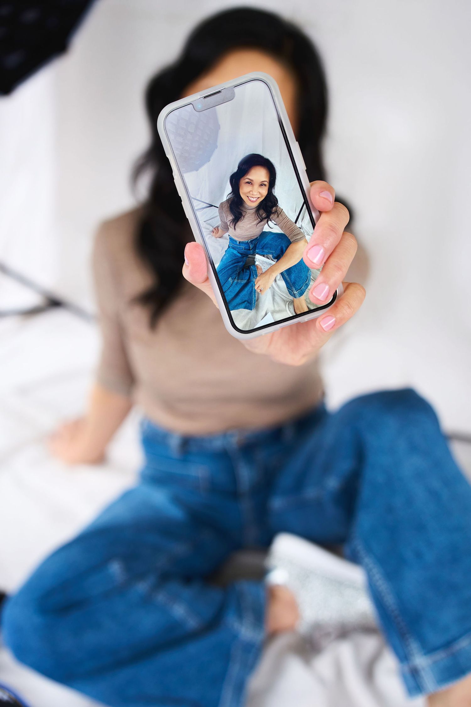 Woman holding smartphone displaying her photo on screen while sitting on bed in jeans and beige top.