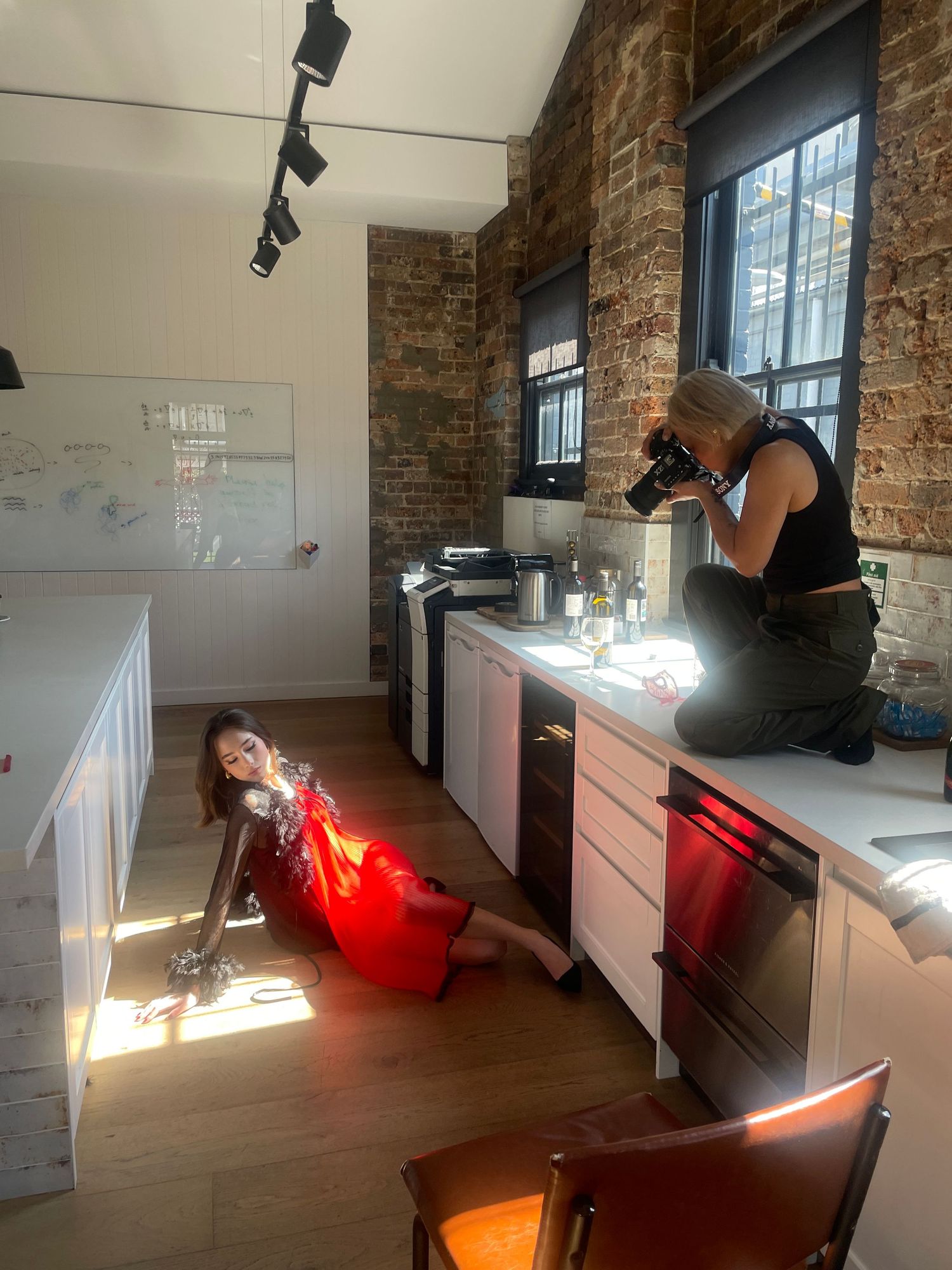 Photographer crouches on a kitchen counter shooting a model in a red dress posed on the floor in a sunlit brick-walled loft studio.