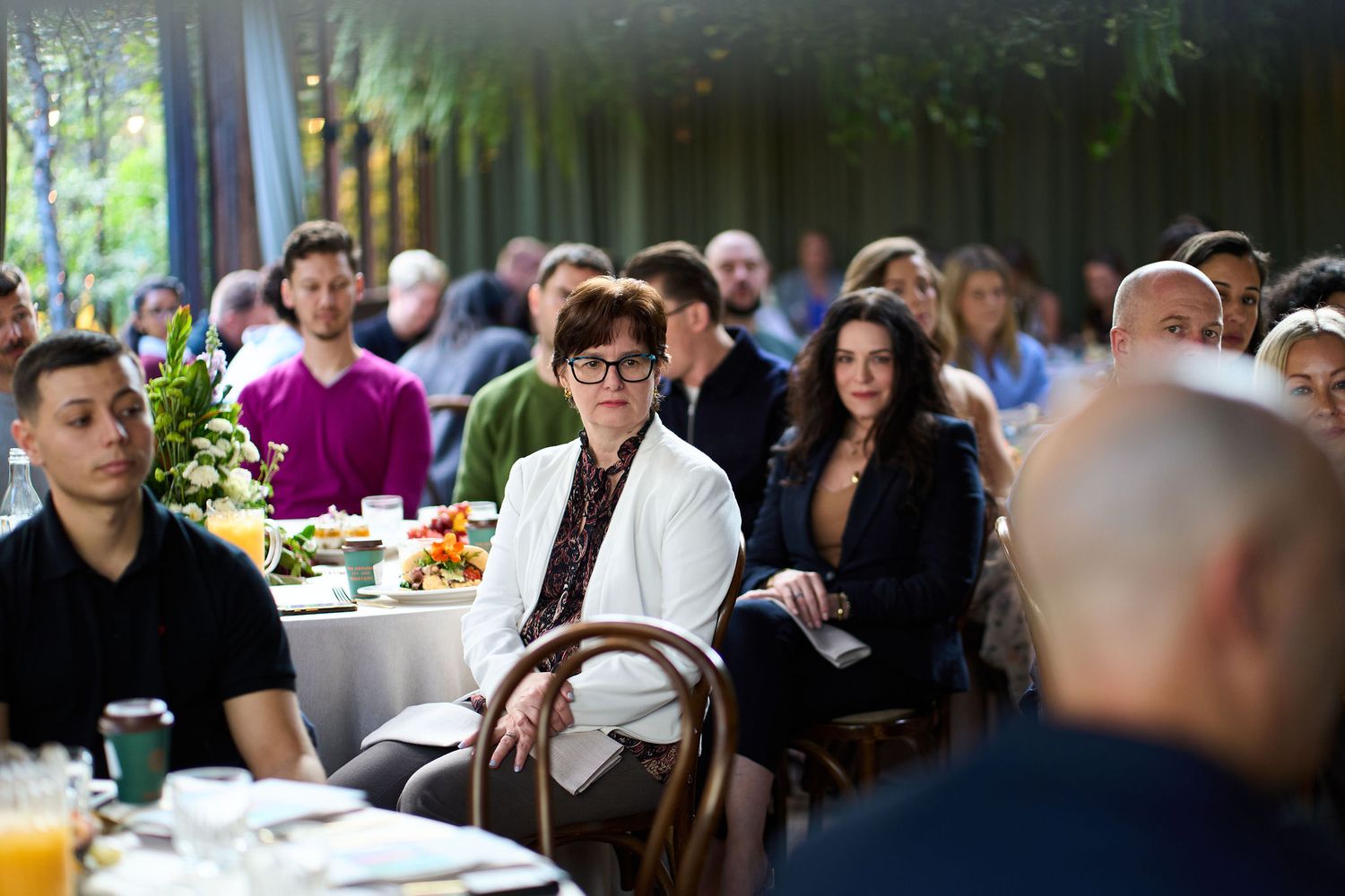 Group of adults seated at tables during an outdoor event or conference, engaged in attentive listening in a garden setting.