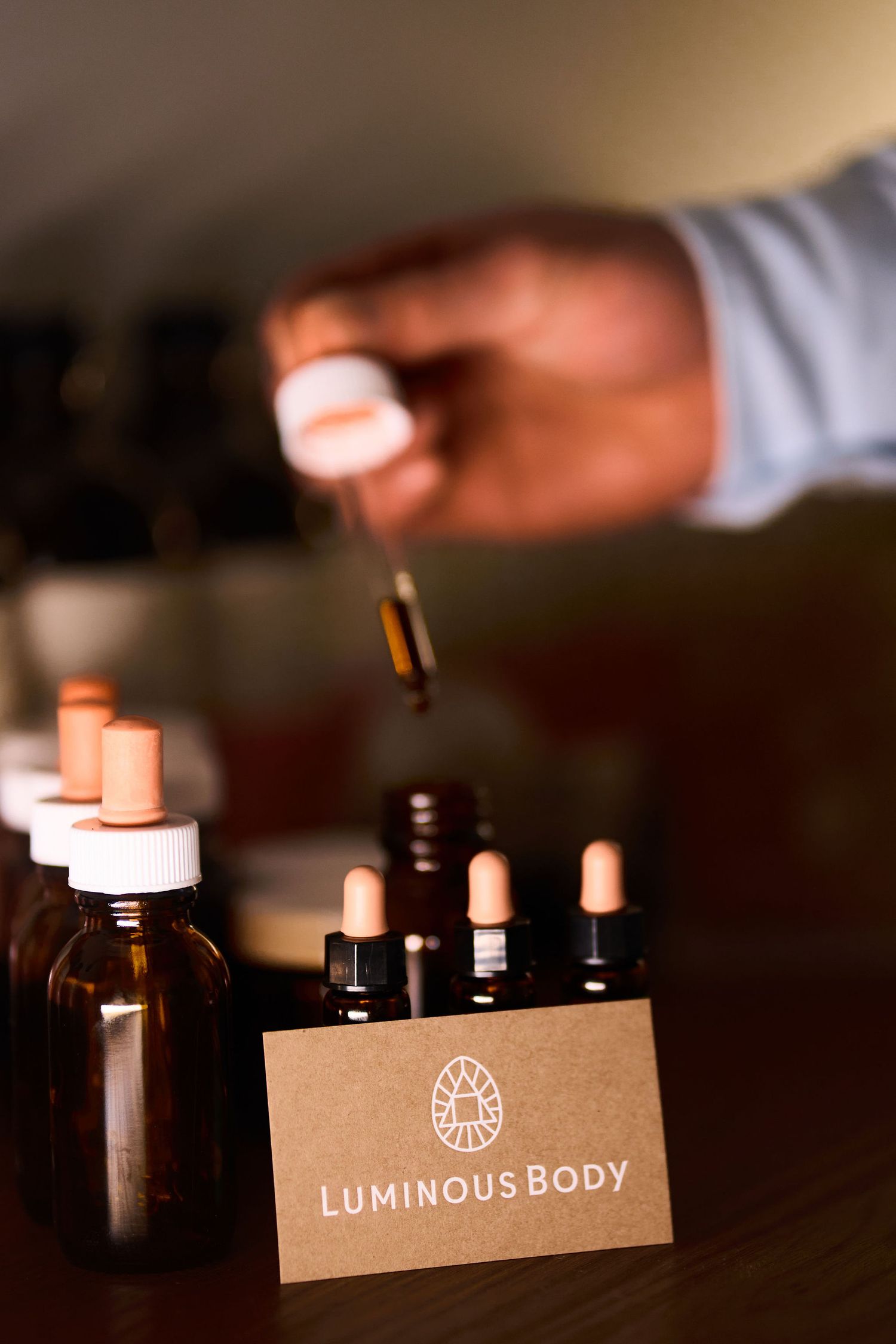 Luminous Body essential oil dropper bottles displayed on a wooden surface with a hand holding a dropper.