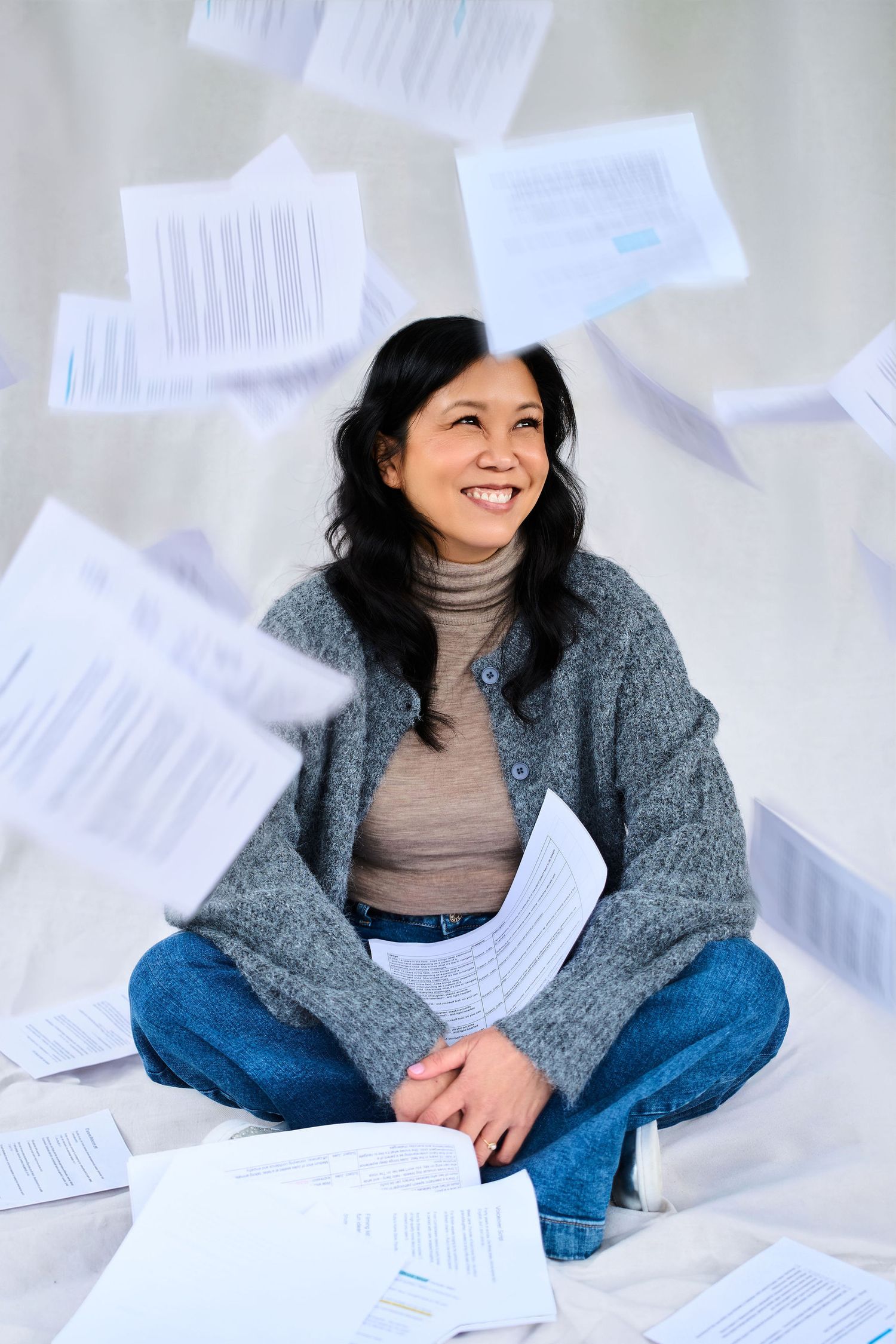 Smiling Asian woman sitting cross-legged on floor with papers flying around her in a bright studio setting.