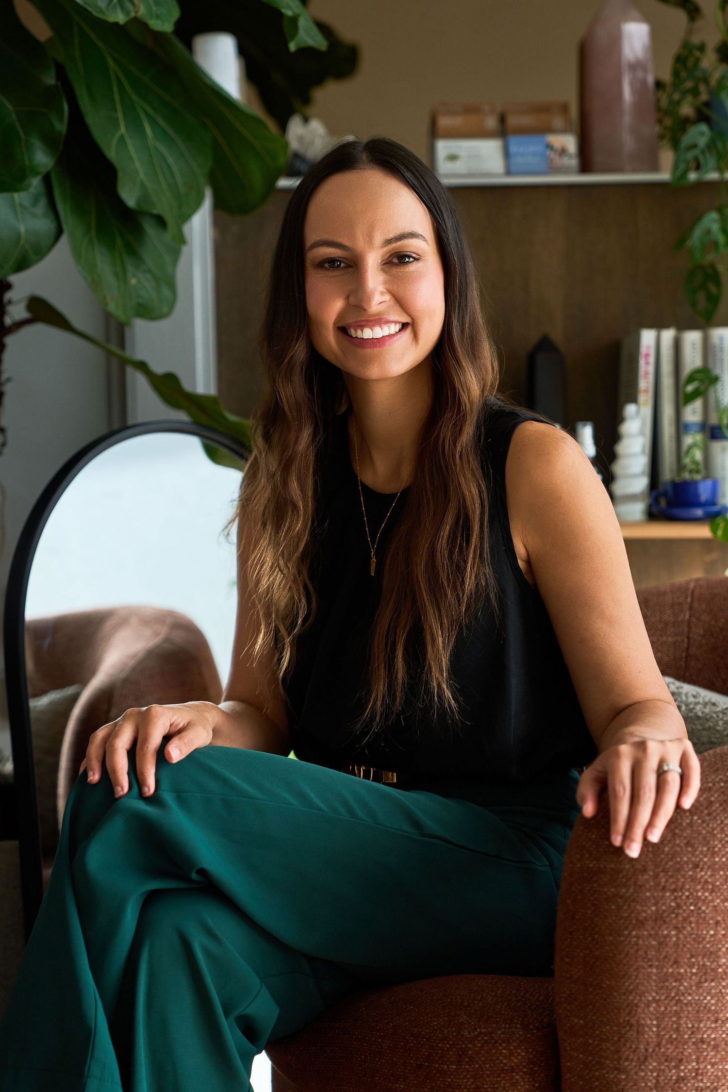 Smiling woman with long brown hair wearing black top and green pants, seated in a modern interior setting.
