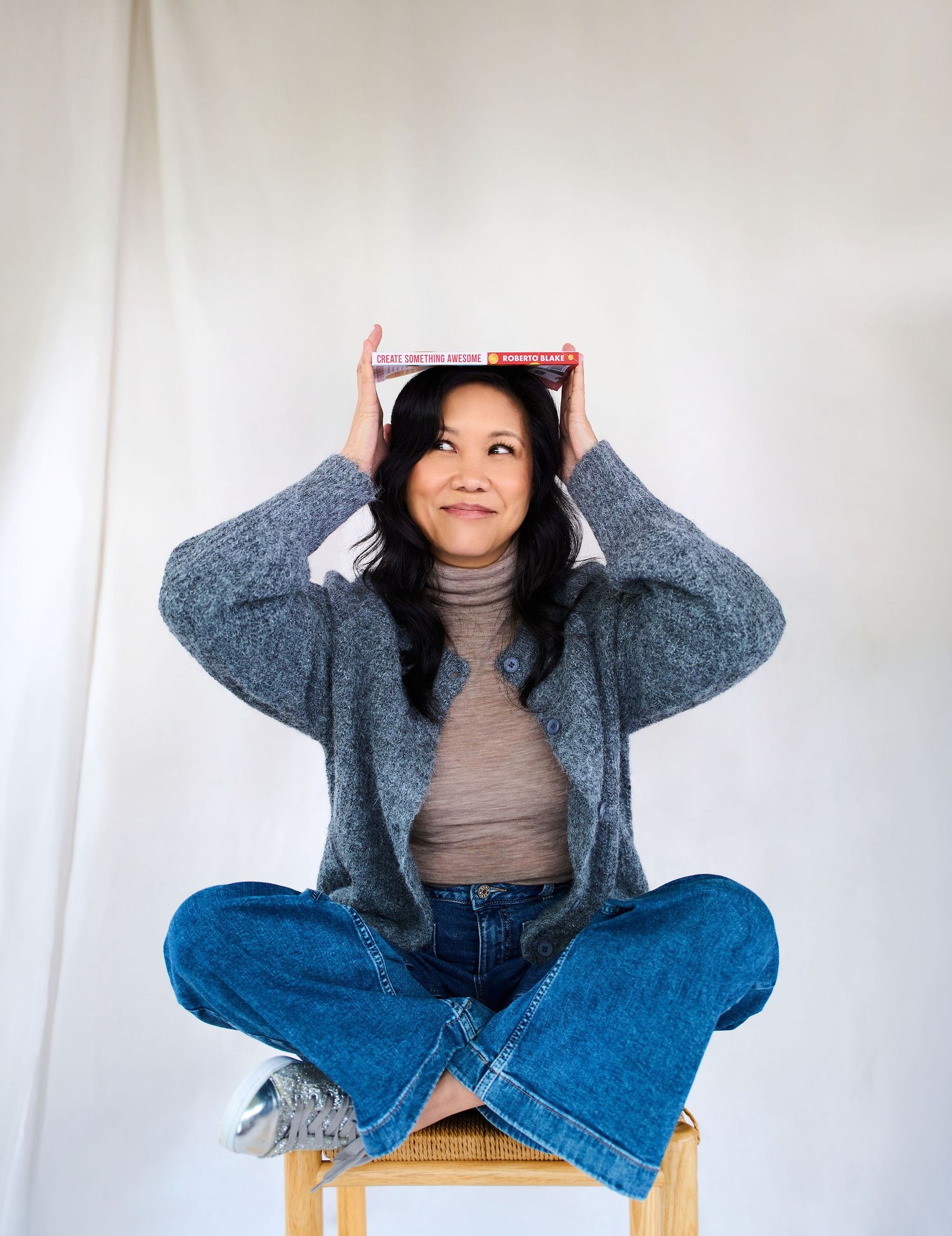 Woman sitting cross-legged on stool balancing a book on her head, wearing gray cardigan and jeans.