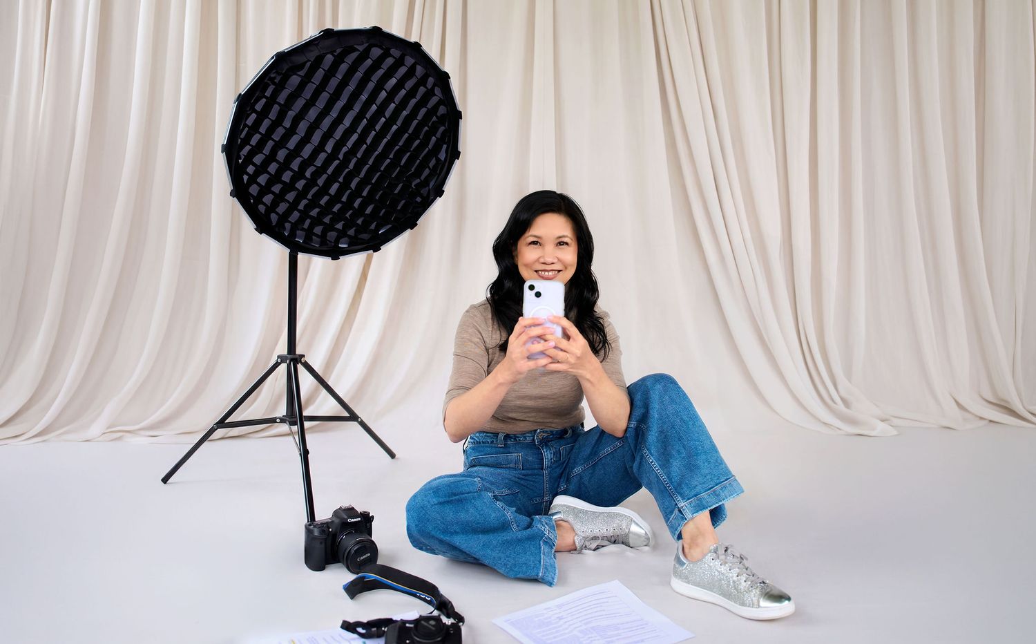 Woman sitting cross-legged with smartphone, photography lighting equipment and camera on white backdrop.